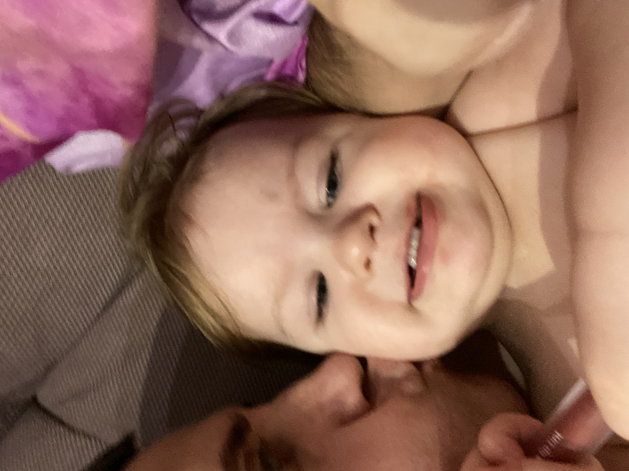 A close-up shot shows a baby with fair skin and light brown hair smiling with visible teeth. The baby's head is slightly tilted. The baby's face is the primary focus. An adult's face is partially visible along the bottom of the frame, appearing to be close to the baby's face. A brown patterned fabric is located on the left side of the image, and lavender and pink fabrics are in the background. The scene is indoors.