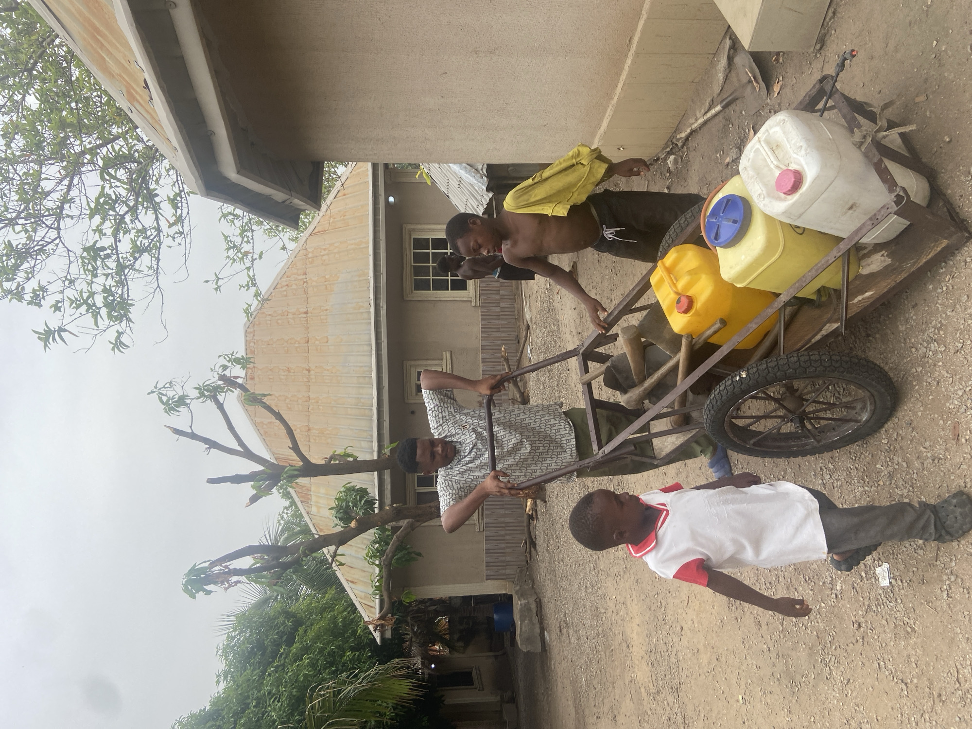 The image, taken in Mararaba, Nigeria, depicts three males and a pushcart loaded with water containers in an outdoor setting. The scene unfolds on what appears to be a dirt or sandy ground, with residential buildings and lush vegetation in the background.
Two of the males are actively engaged with a sturdy, two-wheeled metal cart. One young male, shirtless and wearing dark shorts with a yellow cloth draped over his shoulder, stands near the cart's side, gripping its frame. He is looking down, focused on the task. Another male, dressed in a patterned short-sleeved shirt and dark trousers, stands at the front of the cart, holding its handles as if guiding or pushing it. The cart is laden with three large plastic jerrycans: one white with a pink lid, one yellow with a blue lid, and another yellow one with a red lid, suggesting the transport of water. A small tool handle is also visible on the cart.
A third, younger male, wearing a white polo shirt with red trim and dark pants, stands to the right of the cart, seemingly observing the activity. A partially visible person is also present behind the shirtless male, near a building structure.
The background features buildings with corrugated metal roofs and light-colored concrete walls, indicative of local architecture. Green trees and dense foliage are visible on the left and right sides of the frame. The sky is overcast, providing soft, diffused lighting with no harsh shadows, suggesting either an overcast day or early morning/late afternoon. No discernible text is visible on any objects or clothing. The image captures a moment of daily labor, likely involving water collection or distribution within the community.