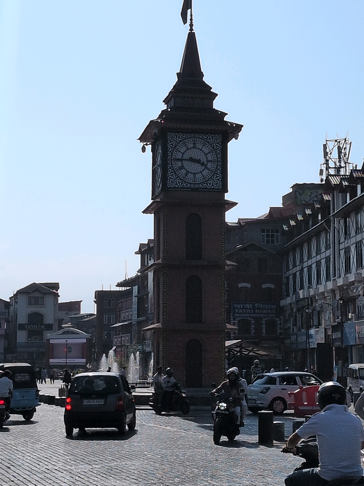 The image depicts a street scene in Srinagar, India, prominently featuring the historic Ghanta Ghar, a clock tower. The tower, made of brick and adorned with intricate clock faces on each side, stands tall against a clear blue sky. Surrounding the tower are multi-story buildings with shops and businesses. In the foreground, a paved street is filled with various vehicles including cars, motorcycles, and a three-wheeled auto-rickshaw. Several people are visible, either riding these vehicles or walking. The atmosphere appears to be a busy urban environment, possibly a market or a central square, with a fountain visible in the background. The architecture of the buildings suggests a blend of traditional and possibly colonial influences.