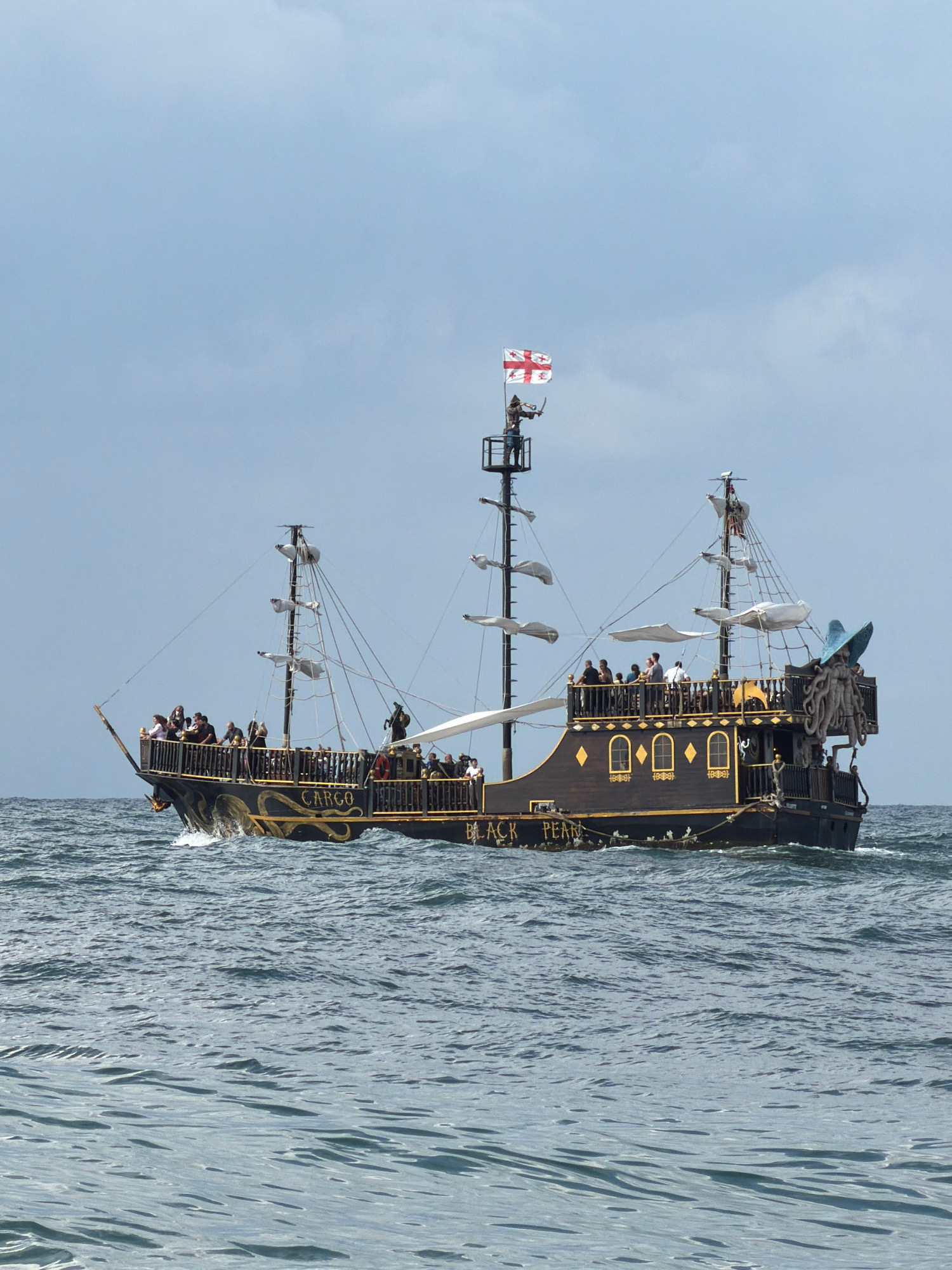 The image shows a pirate-themed ship named "Black Pearl" sailing on the sea, with the city of Batumi, Georgia, visible in the background. The ship is black with gold accents and features multiple masts, sails, and a deck crowded with people. A figure dressed as a pirate stands at the top of the main mast, holding a sword. The ship also has a prominent Kraken-like figurehead on its stern. The sea is choppy, with waves indicating movement. The sky is overcast with light blue and white clouds. People are visible on the deck of the ship, looking out at the sea and interacting with each other. The ship appears to be a tourist attraction or part of an event.