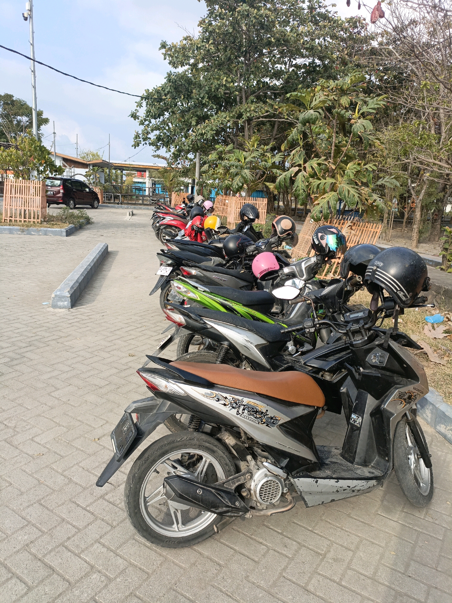 A line of parked motorcycles with helmets on their handlebars are visible in a paved parking area. The motorcycles are of various colors, including black, red, and green. In the background, there is a dark red car parked, along with some trees and a building. The scene appears to be in Bulukumba, Indonesia, given the presence of the motorcycles and the tropical foliage. The lighting suggests it is daytime.