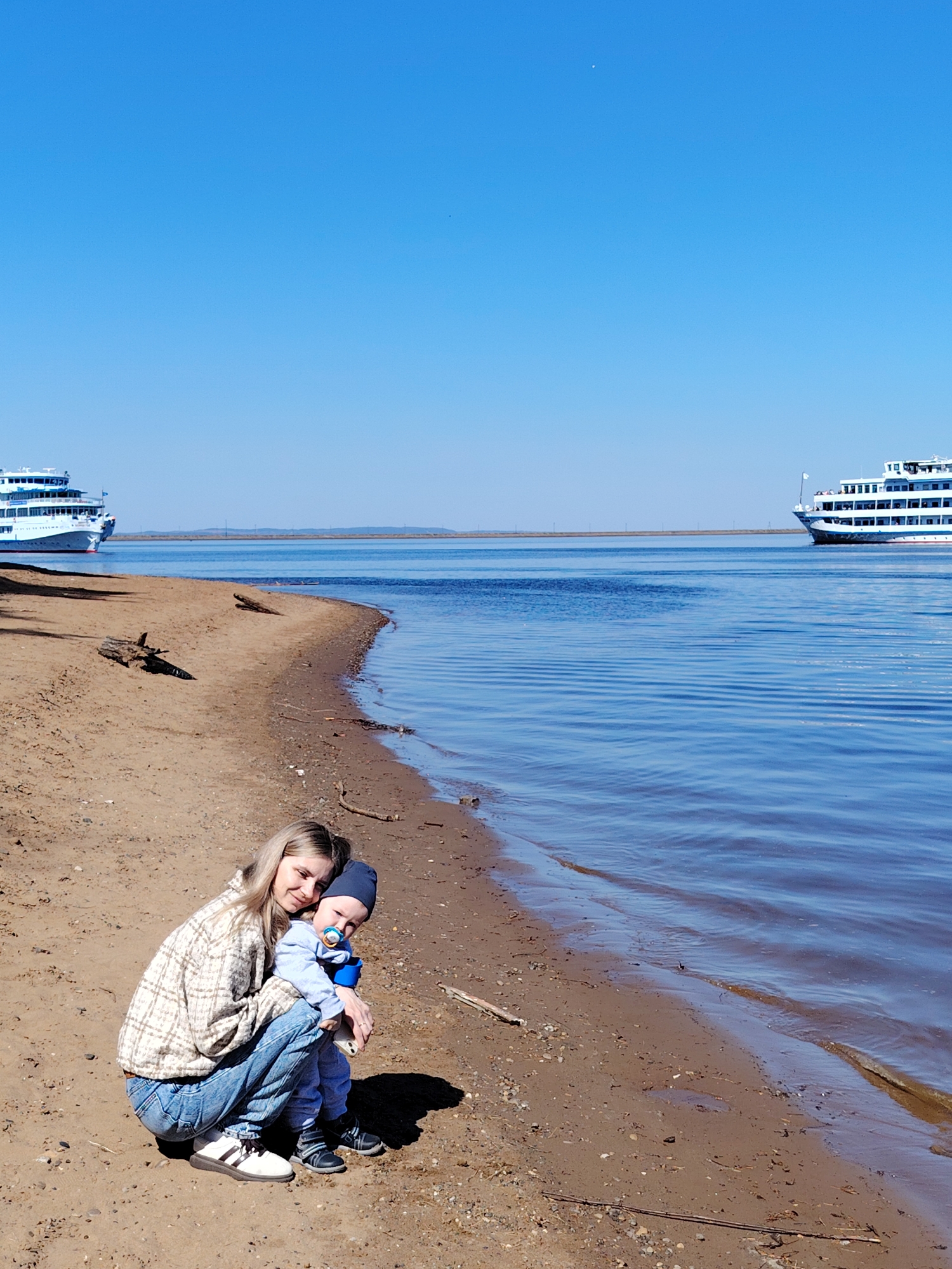 A young woman and toddler are crouched on a sandy beach along the shoreline of the Kama River in Chaikovsky, Russia. The woman, wearing a plaid jacket and jeans, embraces the child, who has a pacifier and wears a blue hat. Calm water stretches to the horizon under a cloudless, blue sky. Two large passenger cruise ships are visible on the river, one to the left and one to the right of the frame. Driftwood and small debris litter the beach.