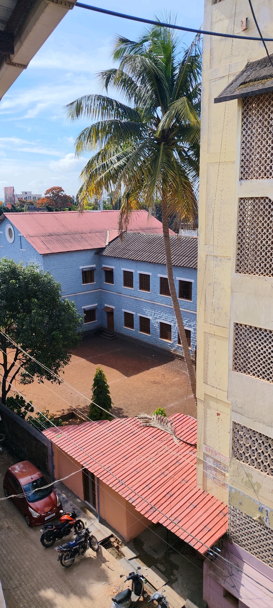 The image captures a scene in Belagavi, India, featuring a light yellow, multi-story building with decorative latticed windows on the right side, adjacent to a blue, two-story building with a red tiled roof. A prominent palm tree stands between the two buildings, its fronds extending into the sky. The blue building has several windows with dark frames and awnings. In front of the blue building, a dirt courtyard is visible with a small, cone-shaped evergreen tree. To the left of the courtyard, a large green tree with dark bark is present. In the foreground, a section of a pink building with a red tiled roof is visible, where a red car and several motorcycles are parked on a paved surface. The overall environment appears to be a school or institutional campus, given the architectural style and open courtyard. The sky is blue with scattered white clouds, indicating daytime.
