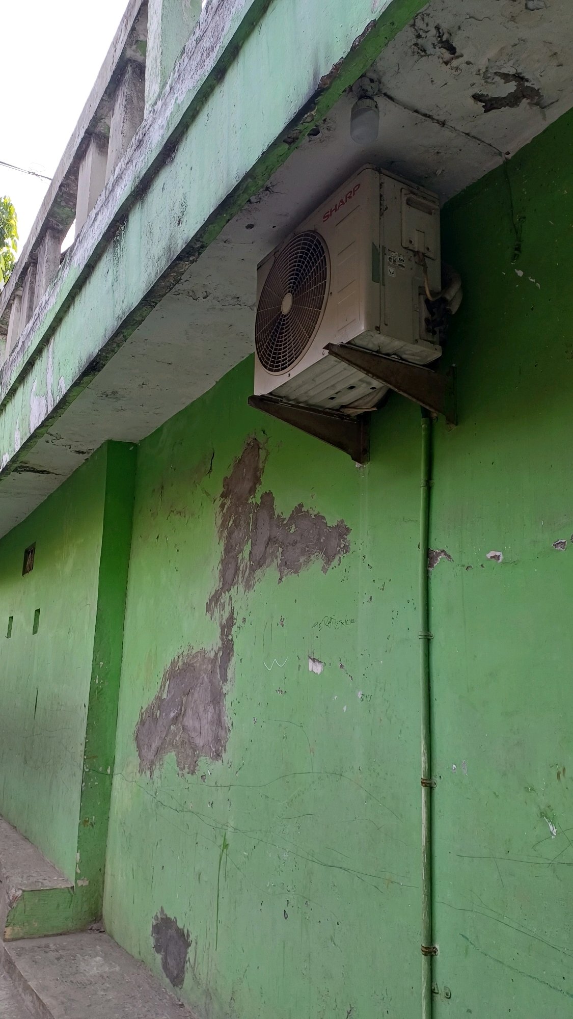 An outdoor, eye-level, slightly angled medium shot captures the exterior of a weathered green wall with patches of exposed concrete. Above the wall, a concrete structure with a light green painted railing extends horizontally, showing signs of peeling paint and water damage. Mounted on the underside of this structure, and partially obscuring the view, is an off-white outdoor air conditioning unit, a "Sharp" model. It is affixed to dark metal brackets and appears to have a cat curled up on top of it. To the right of the air conditioning unit, a green pipe runs vertically down the wall, secured by two metal clamps. The green wall itself exhibits significant discoloration and texture variations, with darker, rougher patches of what appears to be plaster or concrete showing through the green paint. Faint scratches and lines are visible on the surface of the green paint. The lighting suggests it is daytime, with the sky visible in the upper left corner.
