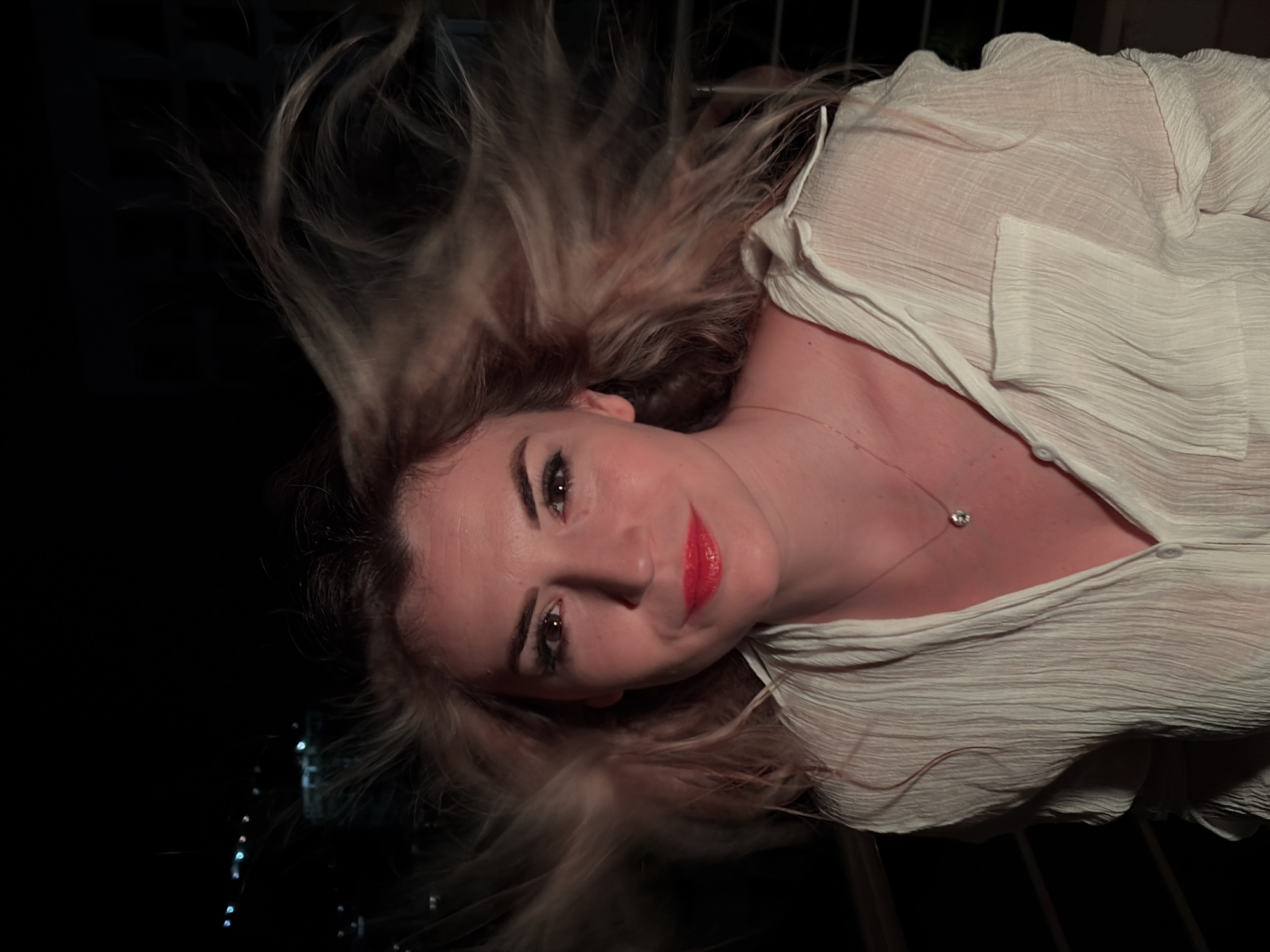 A fair-skinned woman is depicted in a chest-up portrait, captured outdoors at night in the city of Budva, Montenegro. Her blonde hair is centrally parted and dramatically blown upwards and outwards, indicating movement or wind. She features dark, arched eyebrows, visible black eyeliner, and bright red lipstick. Her brown eyes are open, fixed on the viewer, and she exhibits a subtle, closed-lip smile. She is dressed in a loose, light-colored, textured long-sleeved shirt with a V-neck, displaying a delicate gold-toned necklace with a small, round, reflective pendant. The background is dark, with blurred points of light, consistent with distant city illumination, visible in the upper left. The subject is distinctly lit, contrasting with the nocturnal environment.