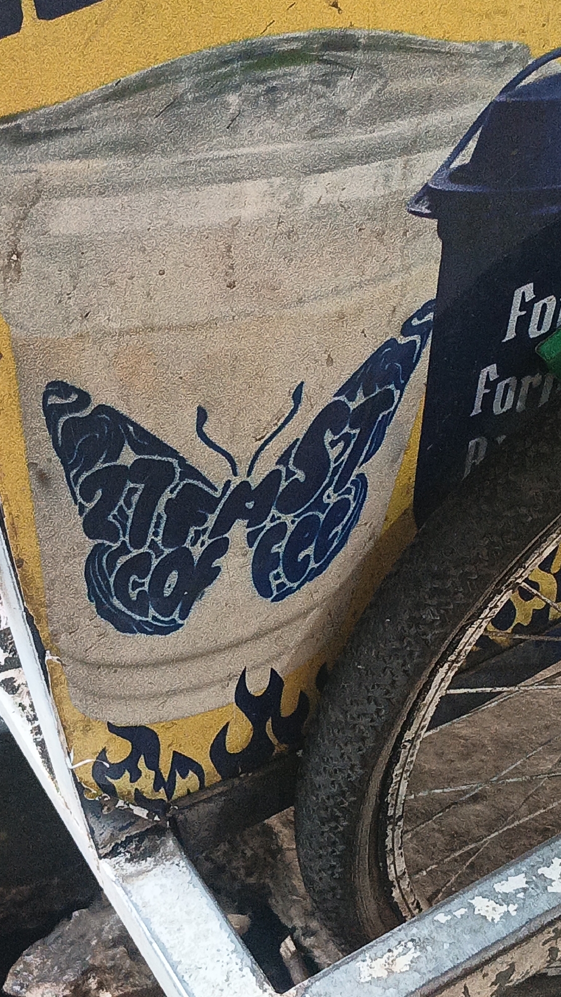 The image shows a close-up view of a textured, weathered surface, likely painted or a decal, featuring a stylized blue butterfly graphic. The butterfly's wings are formed from text, with the left wing appearing to read "27 FF" and the right wing reading "S fee". Below the butterfly, there are stylized black flame graphics on a yellow background. To the right, a portion of a dark blue object, possibly a container or vehicle part, is visible with the white text "For" and "For P" printed on it. Adjacent to this, a bicycle wheel with a worn tire and spokes is prominent. The overall scene suggests an urban environment in Banjarmasin, Indonesia, possibly a street or a market setting, given the commonality of such painted designs on vehicles or stalls in the region.