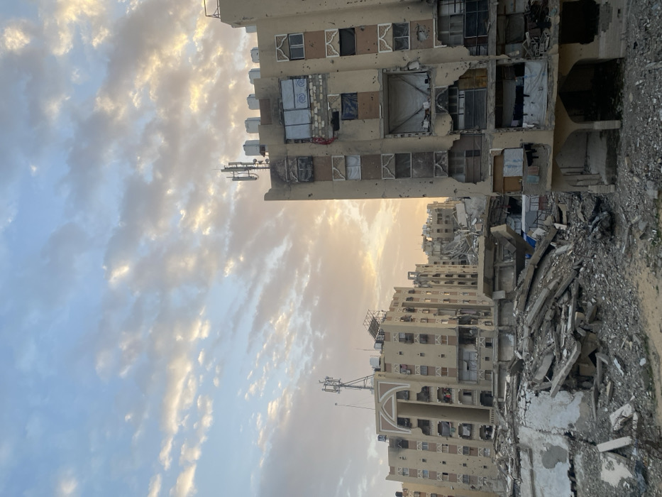 Buildings stand amidst the rubble, bearing scars of war. Windows are shattered, walls crumbled, exposing interiors and hanging garments. The buildings' facades reveal the impact of conflict. Debris litters the ground, a somber testament to destruction. Antennae stand atop the structures, contrasting modern technology with the devastation. The sky glows faintly behind the ravaged buildings.