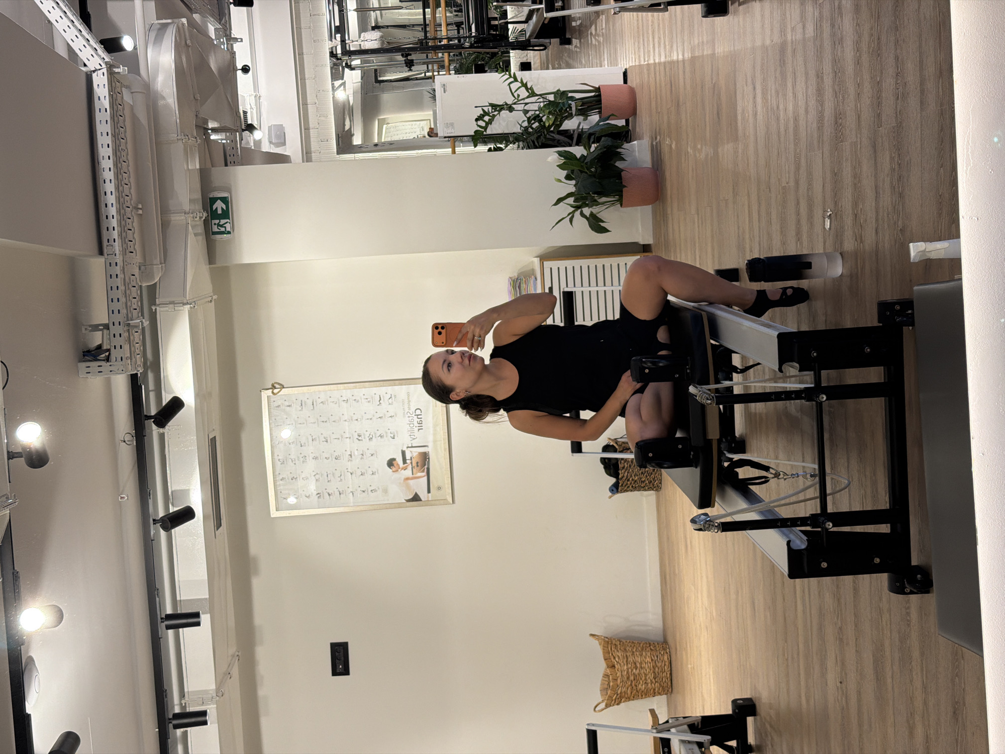 The image depicts an indoor fitness studio, likely a Pilates studio, with an individual performing a workout. The woman, wearing a black tank top and shorts, is seated on a Pilates reformer machine, holding a smartphone to take a mirror selfie. The studio features polished wooden floors, cream-colored walls, and various gym equipment, including reformers and possibly other apparatuses visible in the background. Natural light streams in from windows, supplemented by artificial lighting from track lights on the ceiling. A framed instructional poster hangs on the wall, and several potted plants add a touch of greenery. The overall environment suggests a clean, modern, and well-maintained fitness space. The location of Boreti, Montenegro is not depicted in the image.