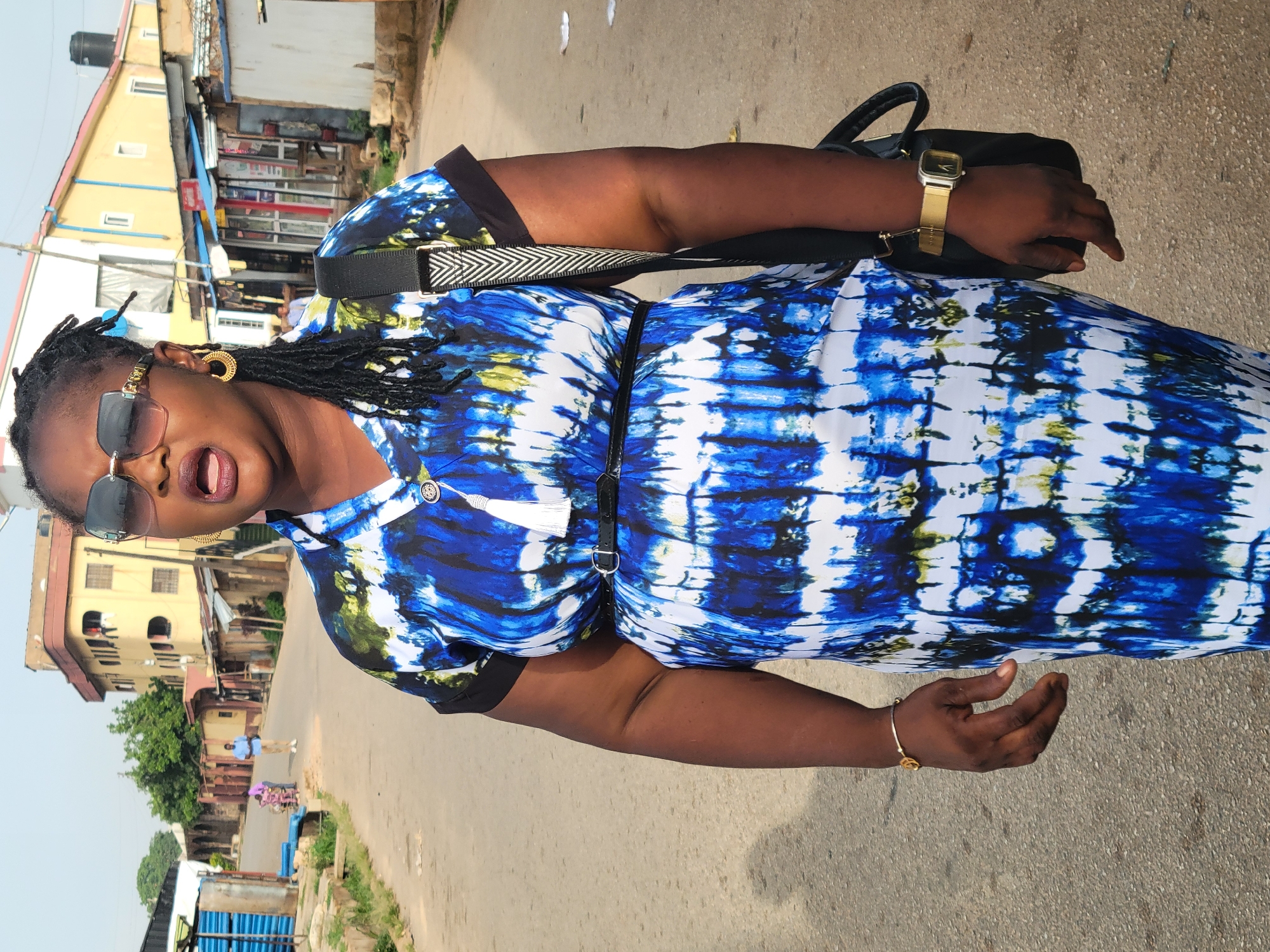 A woman stands on a paved road in Jos, Nigeria. She is wearing a blue and white tie-dye dress with a black belt, black framed sunglasses, and gold jewelry including a watch, bracelet, and earrings. She has dark hair styled in braids. She is facing forward with her mouth open. A black bag is slung over her shoulder. The background includes buildings, trees, and a person in the distance. The sky is visible.