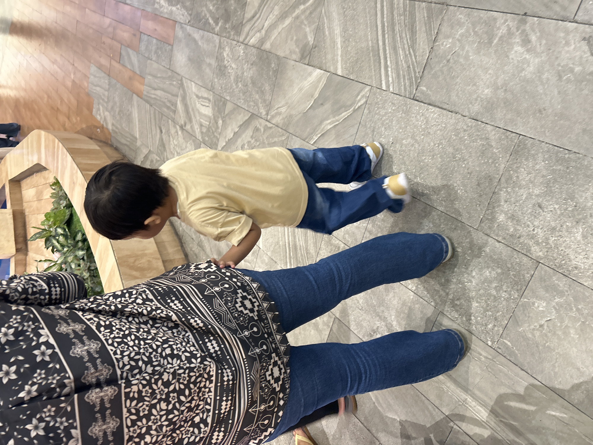 The image shows a young child in a yellow t-shirt and blue jeans walking on a tiled floor. The child is walking away from the camera, towards a planter with greenery. To the left of the child is the lower half of an adult wearing a black and white patterned shirt and blue jeans. The adult's left hand is visible, near the child's back. The floor is a light grey tile with darker grey veining, and to the right, there are wooden planks. The scene appears to be in an indoor public space, possibly a shopping mall or a museum, given the architecture and the presence of plants. The lighting suggests an indoor environment with artificial illumination. There are no overt indications of the specific location being in Makassar, Indonesia.