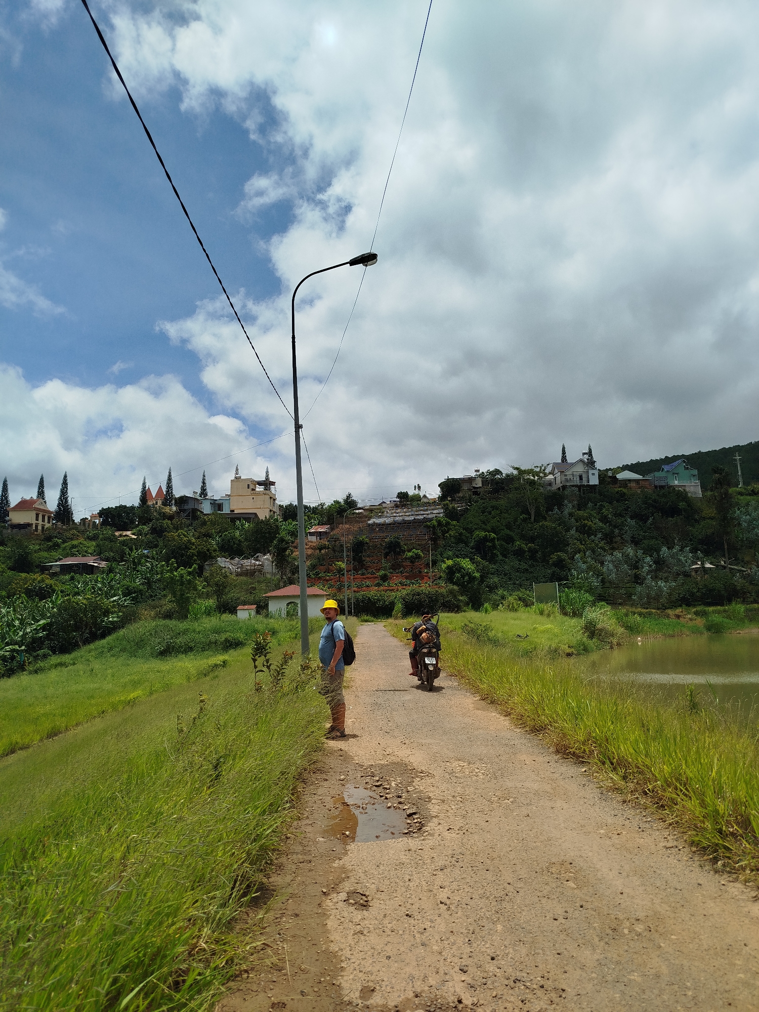 The image depicts a rural scene in Xuân Sơn, Vietnam, on a cloudy day. A dirt road runs horizontally through the frame, bordered by lush green grass on the left and a body of water on the right. In the foreground, a person wearing a blue shirt, khaki shorts, and a yellow hard hat stands on the side of the road, looking towards the camera. They are carrying a black backpack. Further down the road, a motorcycle with two occupants is visible, heading away from the camera. In the background, a hillside is covered in dense vegetation and dotted with several houses, some with distinctive architectural features. A tall lamppost stands near the center of the image, with power lines stretching across the sky. The overall impression is one of a peaceful, everyday moment in a Vietnamese countryside setting.