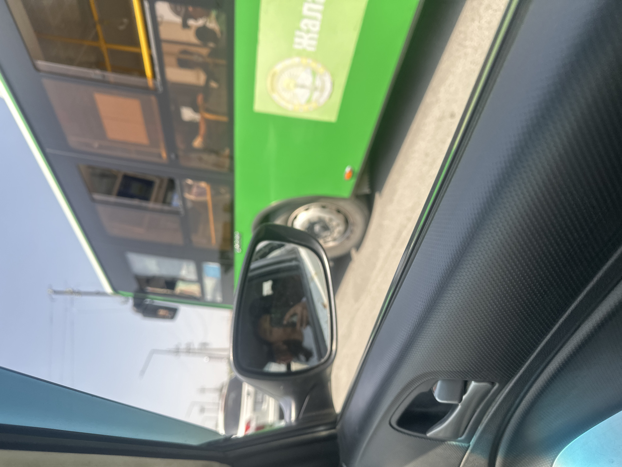 A green bus is driving on a street in Manas, Kyrgyzstan. The photo is taken from inside a car looking out the passenger side window. The bus is large and has several windows. There is a logo on the side of the bus with text. The bus is in motion, and the image is slightly blurred due to the movement. In the rearview mirror, a person is visible taking a selfie. The sky is clear and blue.