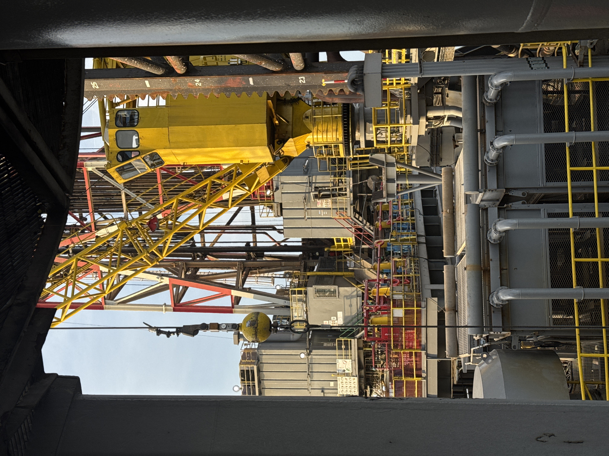 A photograph captures a close-up view of a section of an offshore oil rig, framed by dark grey structures on the left and right. The main subject is the towering yellow drilling derrick with its intricate lattice of red and yellow beams. A yellow control cabin with multiple windows is visible near the top right. Below the derrick, grey industrial modules with prominent yellow railings and staircases are stacked. In the foreground, large grey pipes and ventilation grates are visible. The overall environment suggests an industrial setting on the ocean. The sky is a pale blue. There are no people visible in the image.