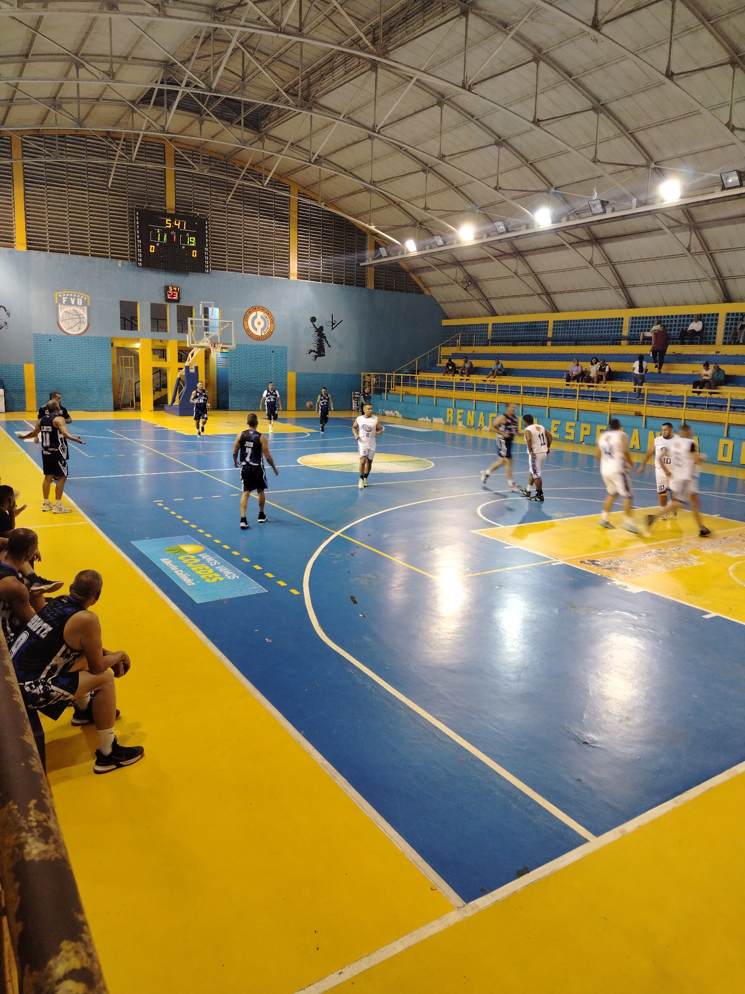 The image shows a basketball game in progress indoors. The court is marked with blue and yellow paint. Players in white and black uniforms are actively playing. Some players are seated on the sideline. The scoreboard indicates a score and time of 5:41. Spectators are seated in the bleachers. Logos are displayed on the wall. Based on the image, "Vamos Vamos Cojedes Alberto Galindez", this photo appears to be taken in Tinaquillo, Venezuela.
