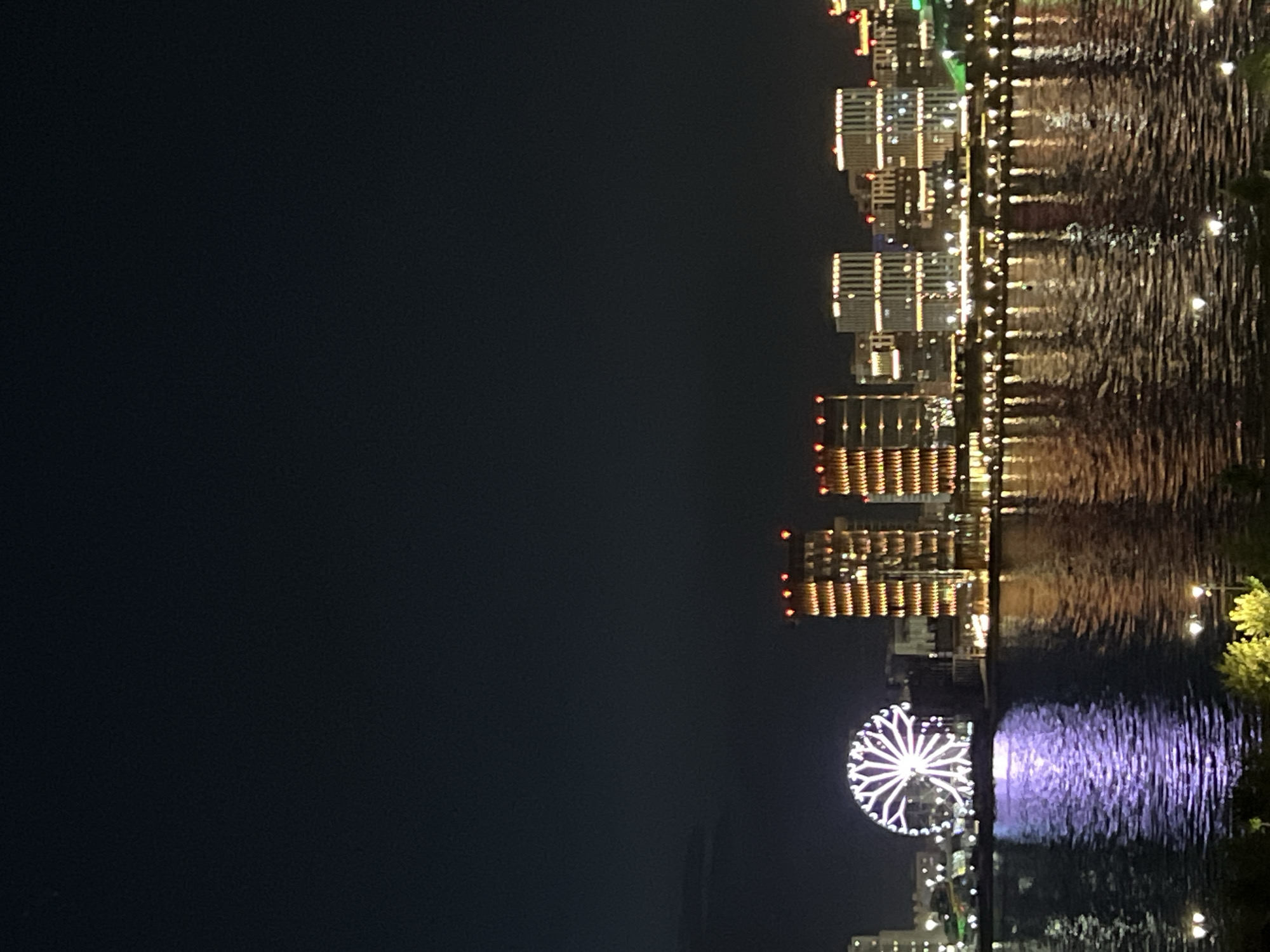 The image captures a night scene in Kazan, Russia, featuring illuminated buildings reflecting in the water. A Ferris wheel with white lights stands out, its reflection adding to the visual effect. The city skyline is composed of multiple high-rise structures with lights on. The water surface shows ripple patterns from the reflections. The sky is dark, contrasting with the city lights.