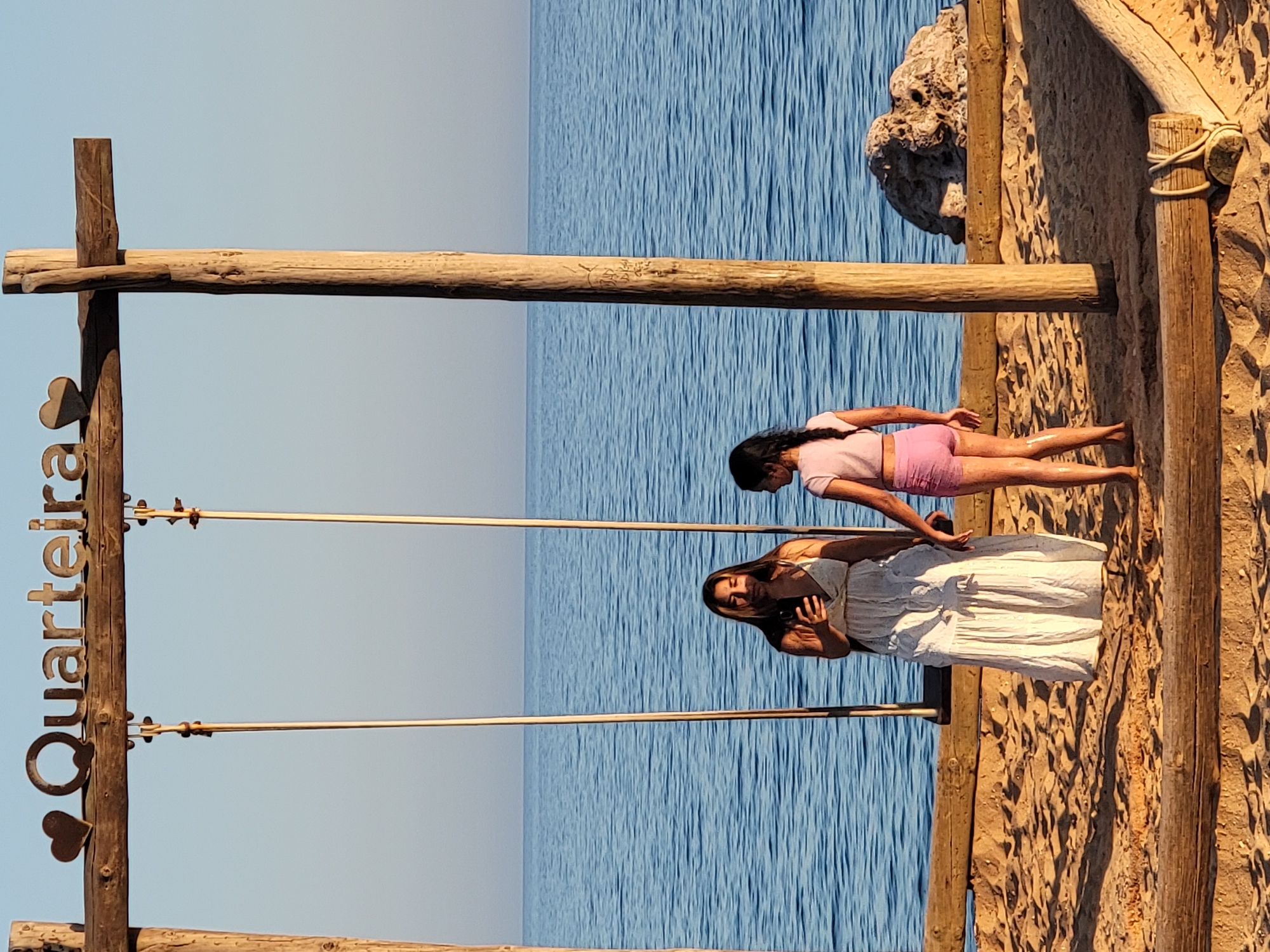 The image captures a serene beach scene in Quarteira, Portugal, with the iconic "Quarteira" sign mounted on a wooden swing set structure overlooking the calm blue ocean. Two individuals, an adult female and a child, are positioned on the sandy beach. The adult, wearing a flowing white dress, is holding a smartphone and appears to be taking a picture or video. The child, dressed in pink shorts and a t-shirt, stands beside her, facing the ocean. The sunlight casts long shadows on the sand, indicating it is either early morning or late afternoon. A large rock formation is visible in the background on the right side of the frame. The overall atmosphere is peaceful and idyllic.