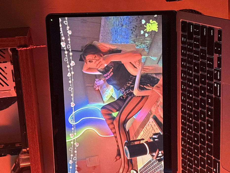 The image displays a close-up of a silver or space grey laptop, likely a MacBook, situated in a dimly lit indoor environment in Yerevan, Armenia. The ambient light casts a prominent reddish-orange hue over the scene.
The laptop screen occupies the majority of the frame, showcasing a still image or live feed. This content depicts a young individual, positioned reclining in a white chair. The individual is wearing a dark t-shirt with light text, a plaid skirt, and black fishnet stockings with horizontal stripes, and is holding a smartphone, seemingly taking a selfie. Their legs are crossed at the ankles and extended leftwards, resting on a surface that appears to be a desk or table. On this surface, a grey keyboard, a black microphone on a stand, and a portion of another laptop are visible.
The background of the screen content reveals a room with a bed to the right, covered by a patterned blue and white bedspread. A prominent, multi-colored neon light fixture, featuring irregular wavy lines in blue, green, yellow, and red, is mounted on the wall behind the individual. Above this, a string of lights with small heart-shaped bulbs is strung across the upper portion of the frame. In the bottom right corner of the displayed image, a cartoonish green creature with two small hearts floating above its head is visible.
Above the laptop screen, a dark wooden shelf or beam is visible, holding several items. These include a clear container, a small metallic component, and the partial structure of what appears to be an electronic device, possibly a computer chassis or internal components. A red braided cord hangs from the shelf on the right. The laptop's keyboard is partially visible in the lower section of the image.