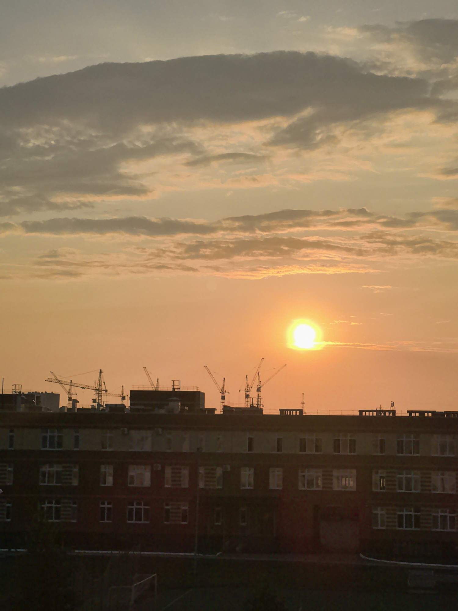 A sunset scene over Froly, Russia, features a large building with multiple floors of windows in the foreground, set against a warm orange and yellow sky. Several construction cranes are visible on top of other buildings in the mid-ground, silhouetted against the sky. The sky is partially obscured by dark, textured clouds in the upper portion of the image, with the bright sun visible as a luminous orb just above the horizon in the right half of the frame. The lower part of the image shows a dark, indistinct area that appears to be ground level or vegetation.