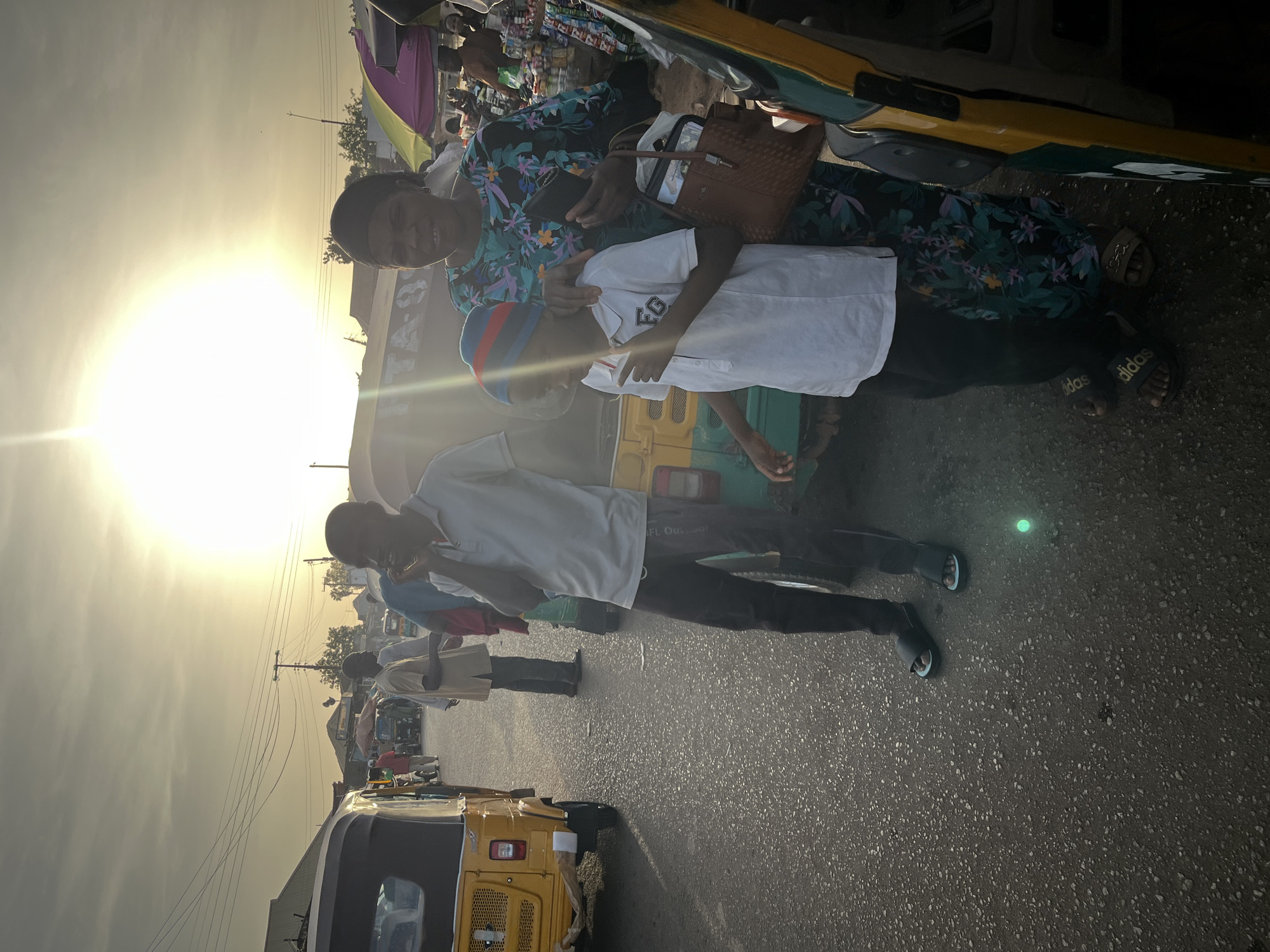 This image captures a bright, bustling street scene in what appears to be a market or commercial area, likely in Jos, Nigeria, given the presence of auto-rickshaws (keke napep). The strong sun flare on the left indicates either early morning or late afternoon, casting long shadows across the asphalt road.
In the foreground, a woman in a vibrant floral dress smiles directly at the camera, holding a dark phone and a brown woven bag. Next to her, a young boy in a white polo shirt and a distinctive blue, red, and white striped hat also looks towards the camera. The boy's shirt has a faint logo that appears to read "FGC" or "EGC." A man in a white polo shirt and dark trousers walks further ahead of them, looking to his left.
The background reveals more activity, with several other people moving about. Dominating the street are yellow and green "keke napeps," which are common in Nigeria. One green keke napep has partial text visible on its side, possibly "BEL OUR F.O." or similar. Stalls and shops line the street further back, displaying various items, suggesting a busy marketplace. Utility poles and power lines crisscross the hazy sky. On the ground, the asphalt road is visible, and at least one person can be seen wearing black Adidas slide sandals. A faint textual element, possibly "0-ALI," is discernible on a structure in the distance. The overall impression is one of everyday life unfolding in a lively urban environment.
