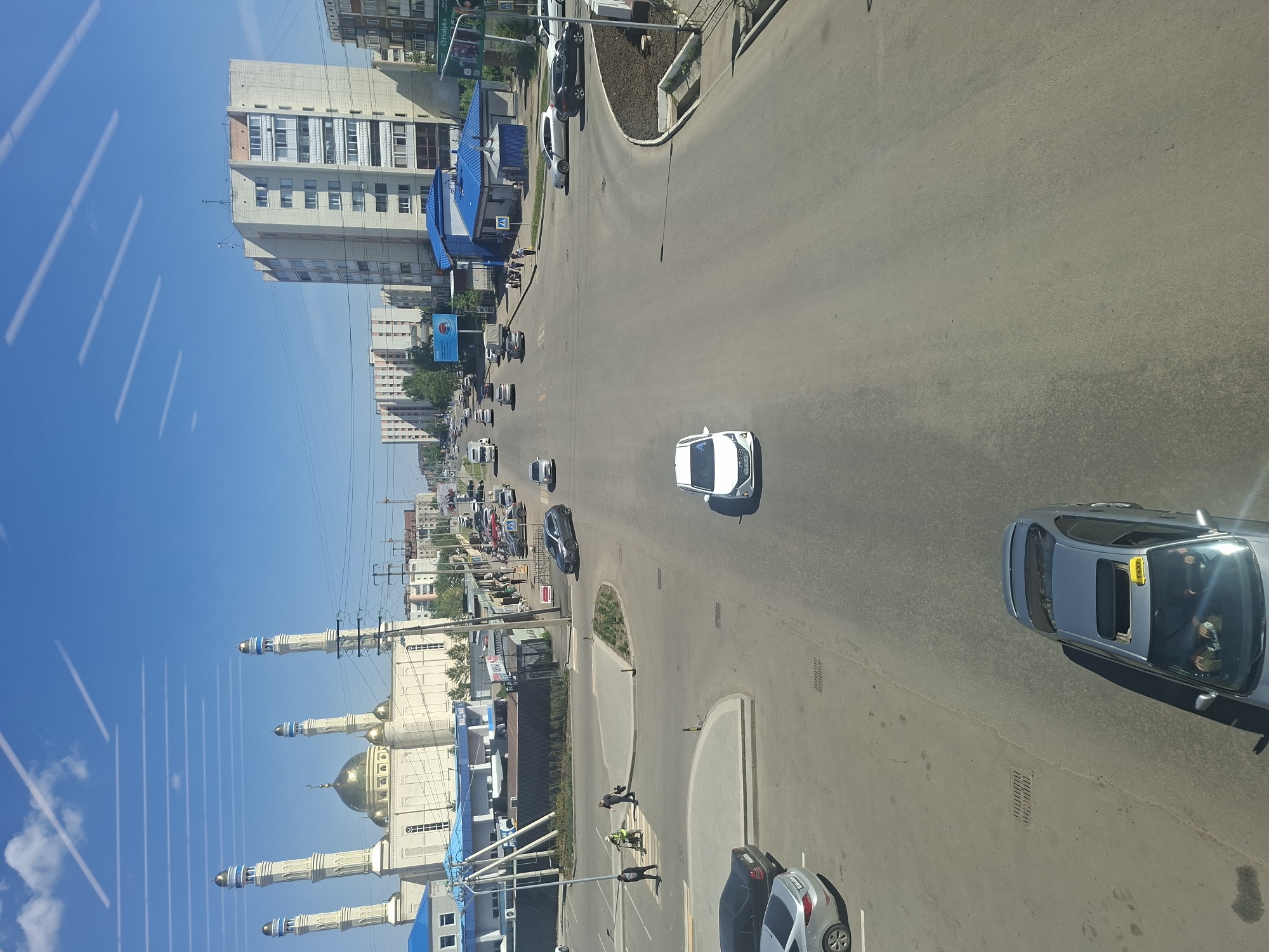 The image depicts a street scene in Kokshetau, Kazakhstan, from an elevated perspective. The Khazret Sultan Mosque is visible on the left side of the image, distinguished by its white minarets and gold dome. A multi-lane road runs through the center, with several vehicles visible, including cars of various colors and sizes. Pedestrians can be seen crossing the street. Buildings of various heights are visible on both sides of the road. The sky is clear and blue.