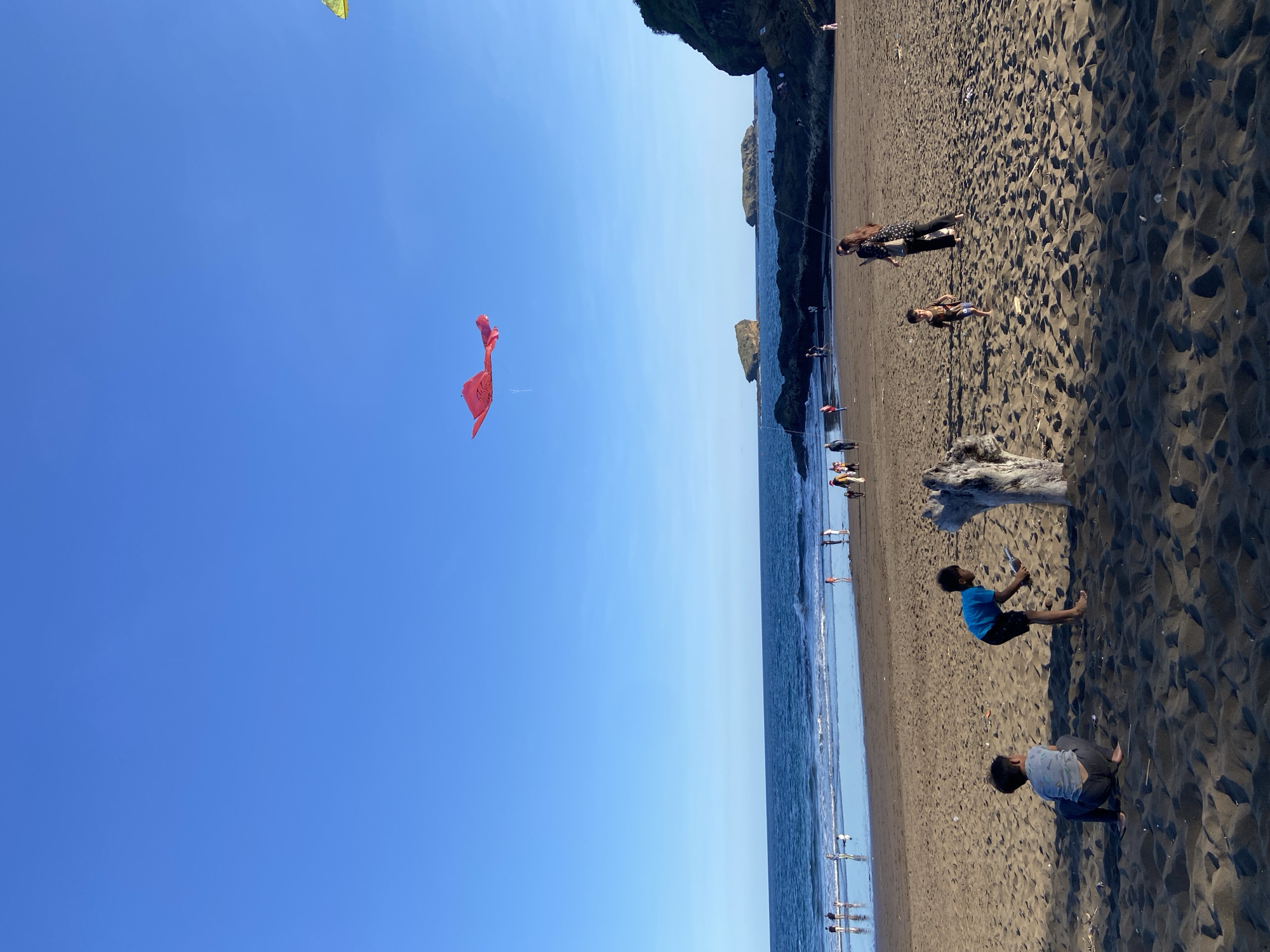 The image shows a beach scene in Unknown, Indonesia. A red kite is flying in a clear blue sky. The beach has dark sand. People are scattered along the shoreline, including some in the water. Two children are playing in the sand near a piece of driftwood. A cliff or rock formation is visible on the right edge of the frame.