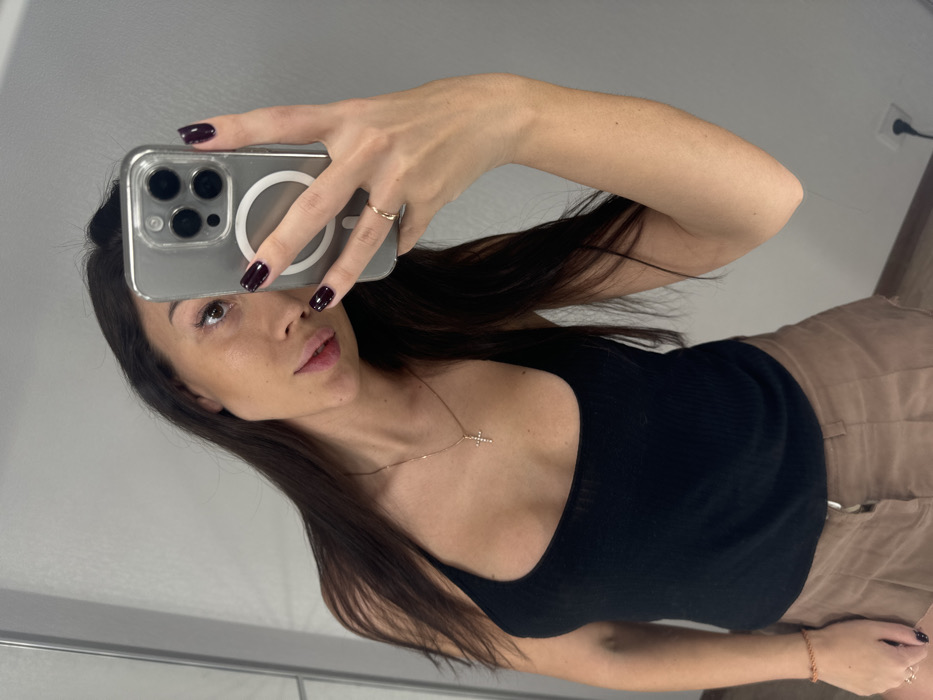 A woman takes a mirror selfie. She holds an iPhone with a clear case in front of her face. She wears a black tank top and khaki shorts. A delicate gold cross necklace adorns her neck. Her long dark hair is parted in the middle. She has dark nail polish and a gold ring on her finger. The background shows a simple wall and a glimpse of an electrical outlet.