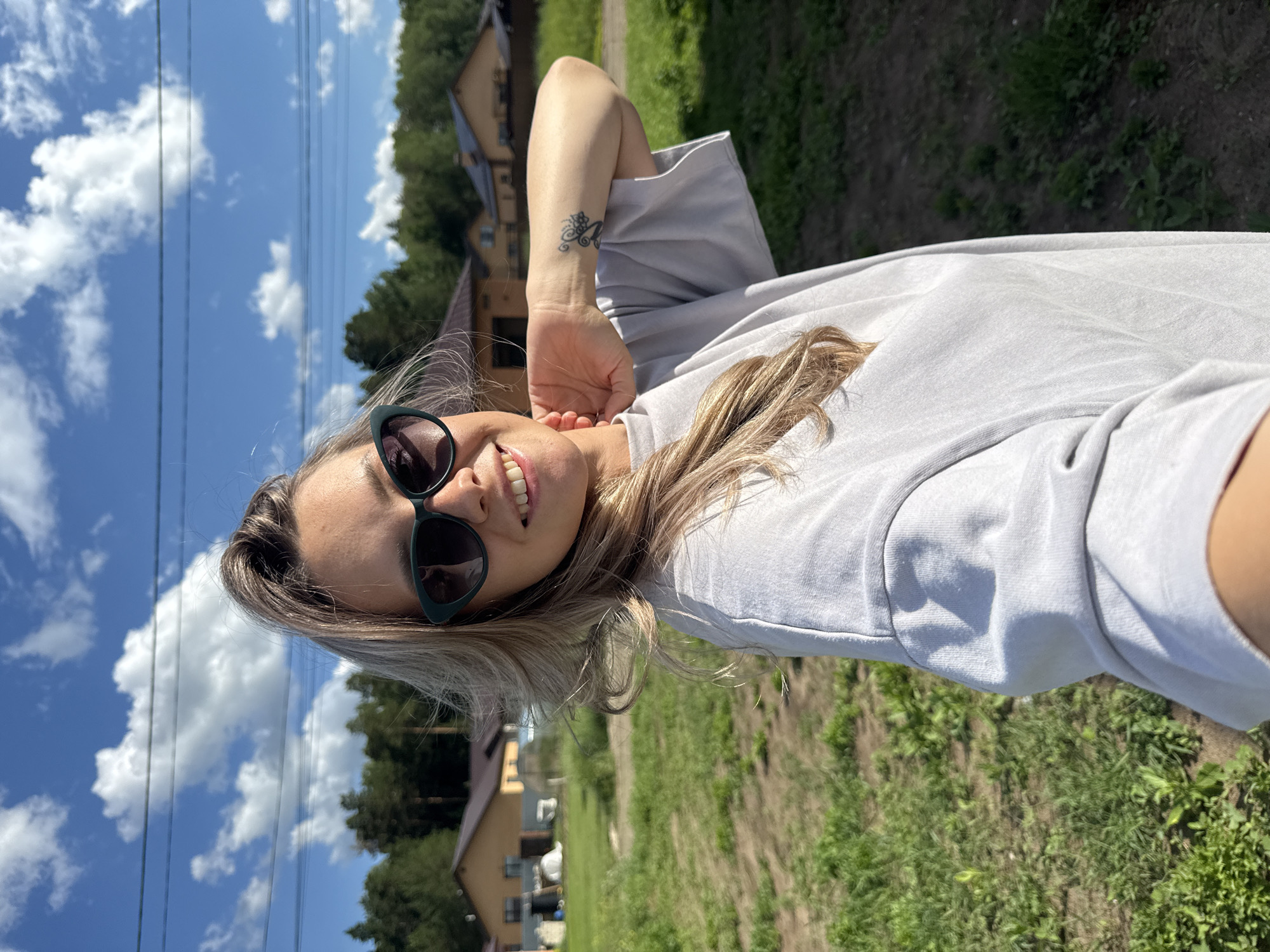 A young woman with blonde hair, wearing sunglasses and a grey t-shirt, smiles at the camera in a selfie. She has a tattoo on her left forearm. The scene is outdoors, with a bright blue sky filled with fluffy white clouds. In the background, there are trees, houses with light brown roofs, and a grassy area. The image appears to be taken on a sunny day, likely in the afternoon. The overall mood is cheerful and relaxed.