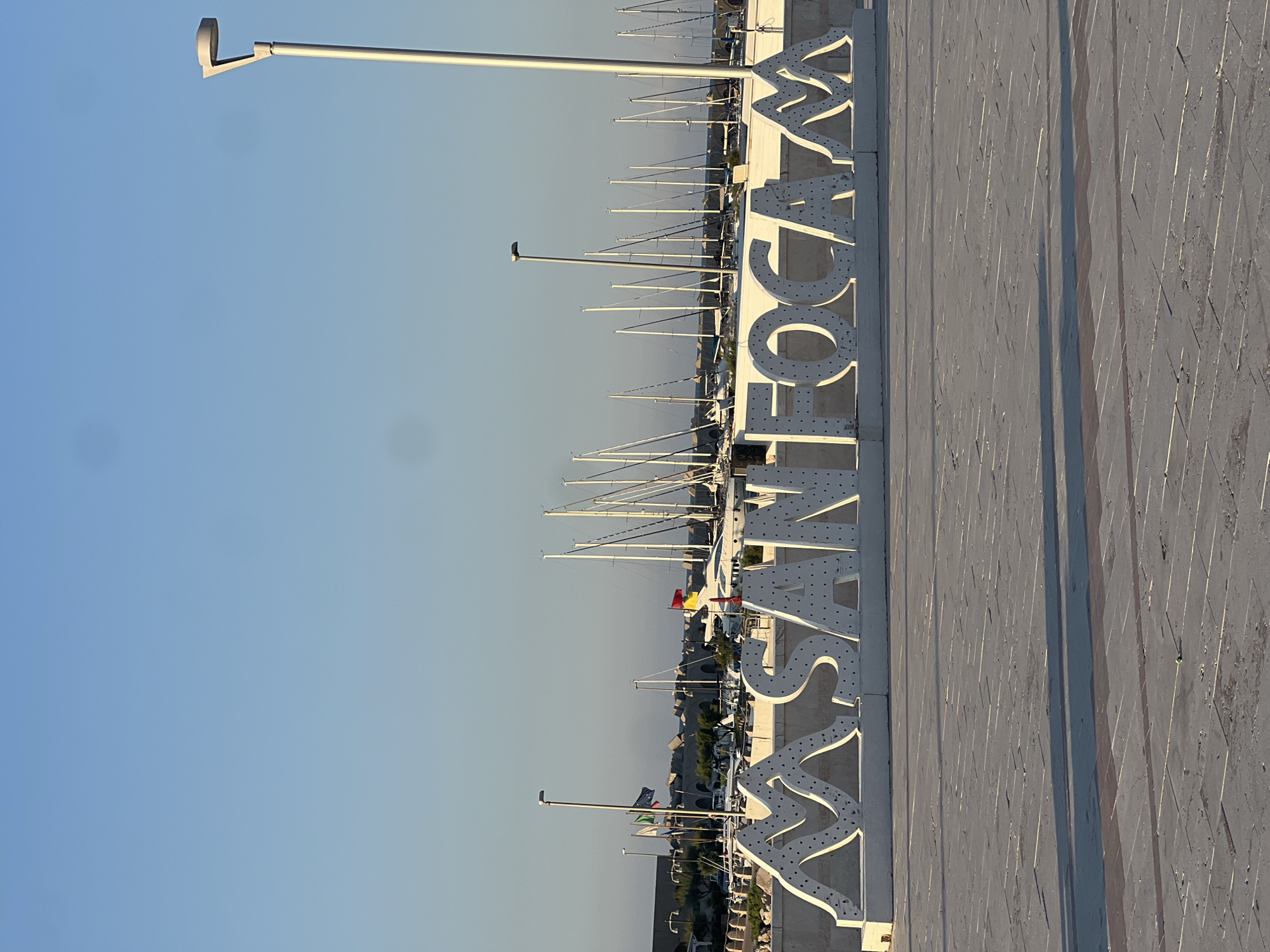 A wide-angle, low-angle, eye-level shot shows a bright, sunny day in the city of San Foca, Italy. The foreground is dominated by a paved, light-colored plaza with several parallel lines and some scattered debris. In the mid-ground, a large, white, three-dimensional sign spelling out "MASANFOCA" is the primary focus. The sign's letters are studded with small, dark circles, possibly for lighting. Behind the sign, a harbor is visible, filled with numerous sailboats with tall, white masts. Some buildings and a sea wall made of concrete blocks are also present in the background. A modern lamppost stands to the right of the frame. The sky is clear and a pale blue, with a slight gradient towards the horizon. There are no people or animals visible in the scene.