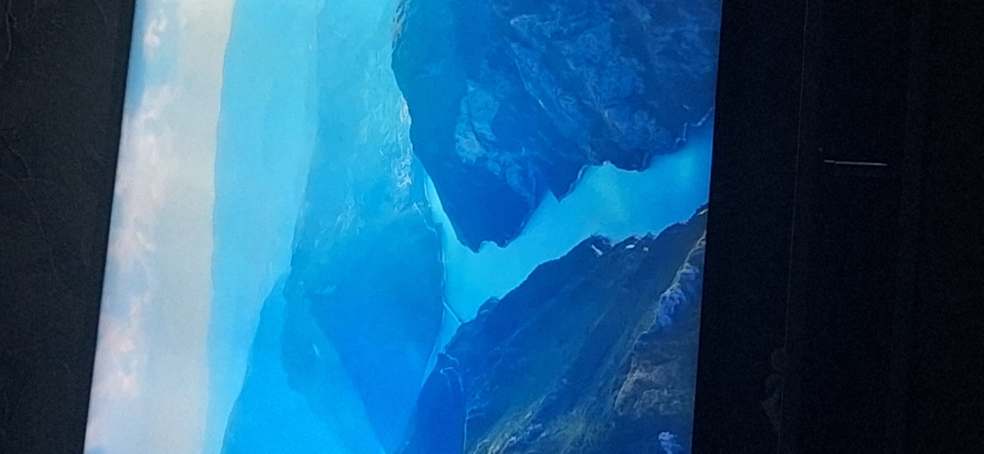 The image shows an aerial view of a fjord-like landscape with a winding body of water surrounded by steep, blue-toned mountains. The sky above is cloudy and hazy, with layers of mountains receding into the distance. There are no people, animals, or man-made structures clearly visible in the immediate landscape. The bottom portion of the image is dark and out of focus, suggesting it may be a photograph of a screen. There is no discernible information in the image that would link it to Bila Tserkva, Ukraine.
