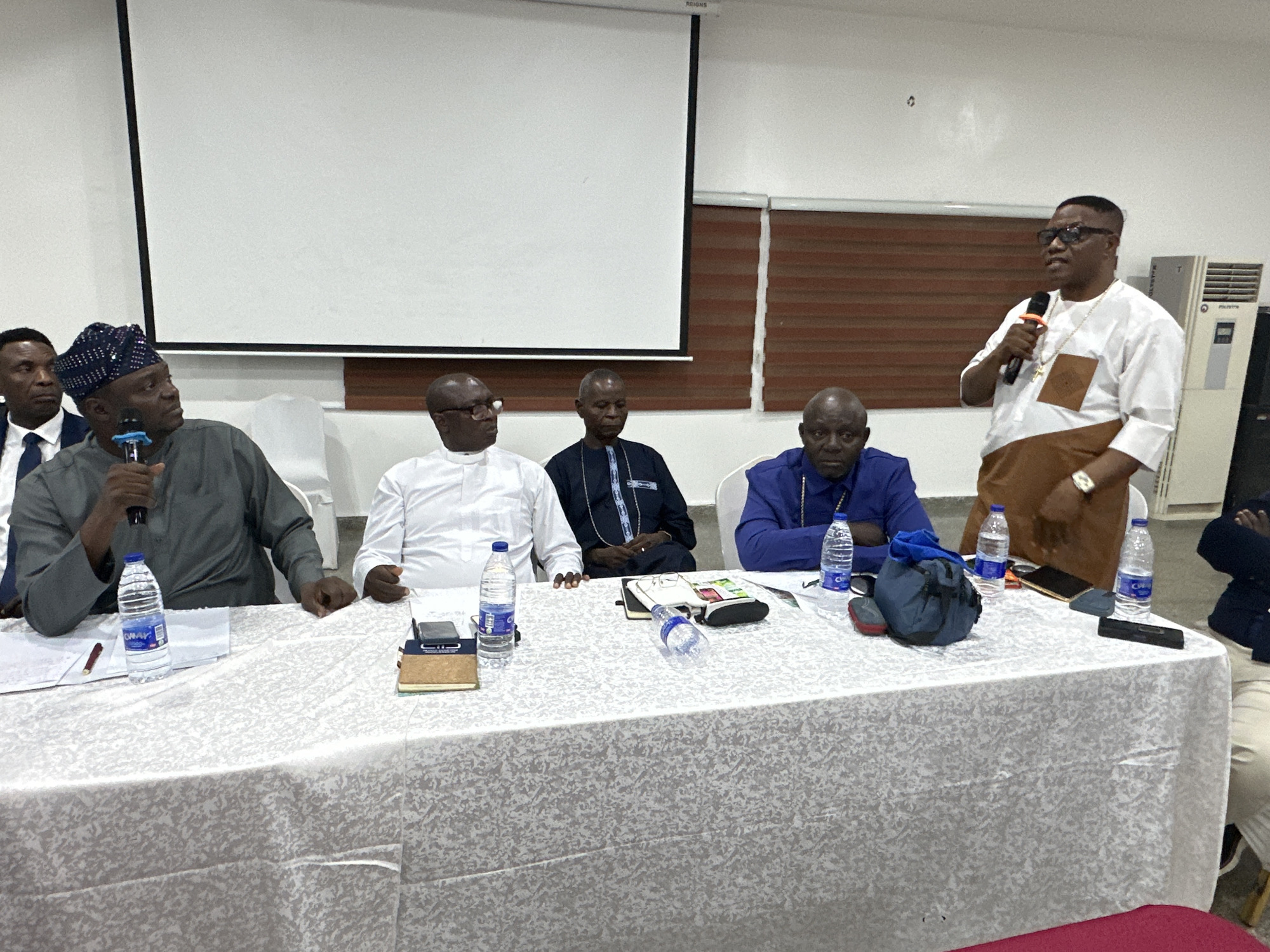 In a meeting room in Abuja, Nigeria, five men are seated around a table covered with a white tablecloth. On the left, a man wearing a dark blue suit and tie is partially visible. Next to him, a man in a grey traditional outfit and a patterned cap holds a microphone and speaks. To his right, a man in a white traditional outfit is seated. Further right, a man in a dark blue traditional outfit is also seated. The furthest man on the right, standing and speaking into a microphone, wears a white and brown traditional outfit, glasses, and gold jewelry. On the table are several bottles of water, notebooks, pens, glasses, a phone, and a small blue bag. The room has a projector screen in the background and a large air conditioning unit on the right.