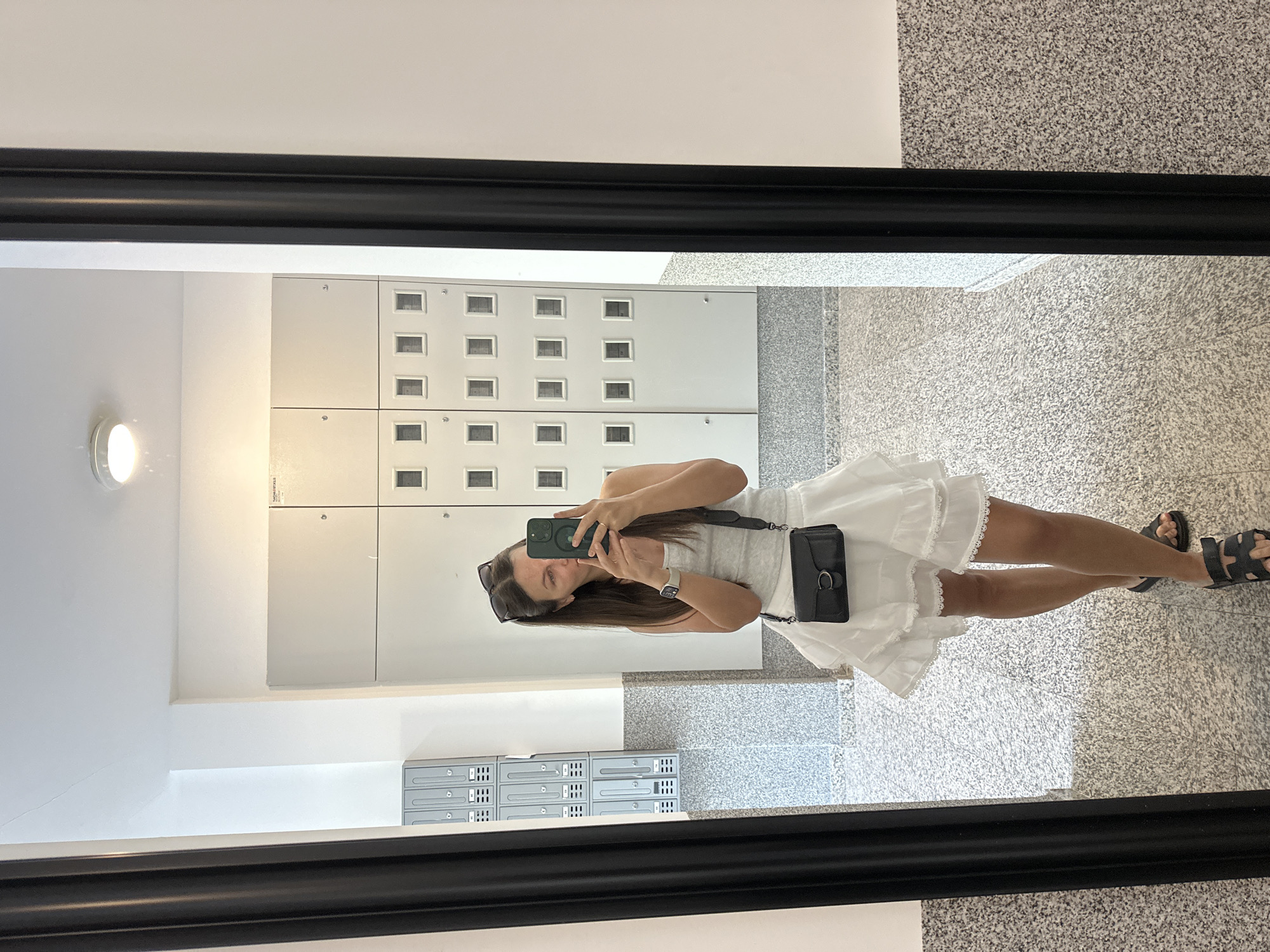 A woman is taking a photo of herself in a mirror. She is wearing a white dress, sunglasses, and black sandals. She is standing in a hallway with gray tile flooring. The wall behind her has white panels with small rectangular windows. A light fixture is visible on the left wall. Mailboxes are attached to the wall in the lower left. The location is an indoor space in Donji Pobori, Montenegro.