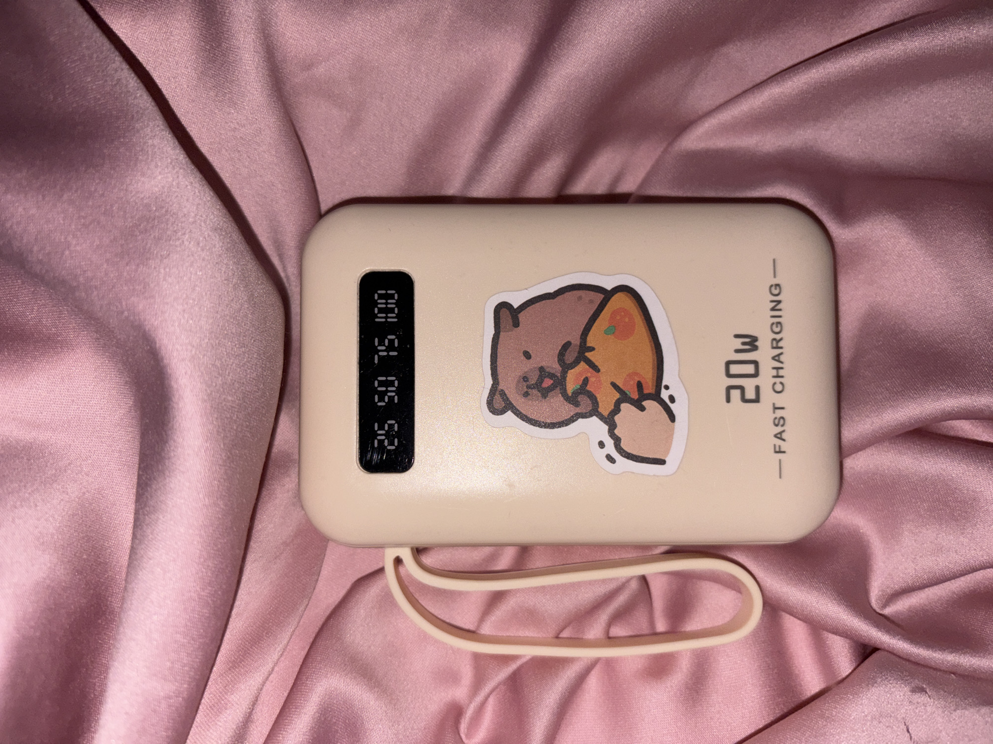 A cream-colored portable charger with a cute sticker of a bear holding a slice of pizza is placed on a pink, satin fabric. The charger has a digital display showing "25 50 75 100" and text that reads "20W - FAST CHARGING". A cream-colored loop is attached to the side of the charger. The pink fabric is draped and wrinkled, creating soft folds and highlights.