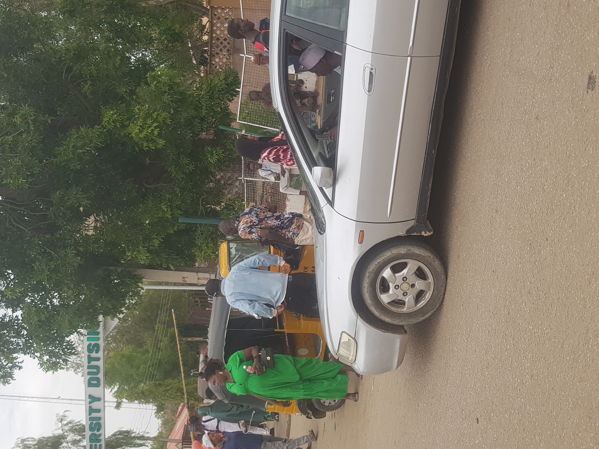 The image depicts a street scene in Dabawa, Nigeria. A silver sedan is parked on the side of the road. A yellow tricycle is adjacent to the sedan. Numerous people are standing near the tricycle and the car. One woman wears a green dress and carries a purse. A sign reading "University Dutsi" is partially visible in the background. Trees and buildings are also visible in the background.