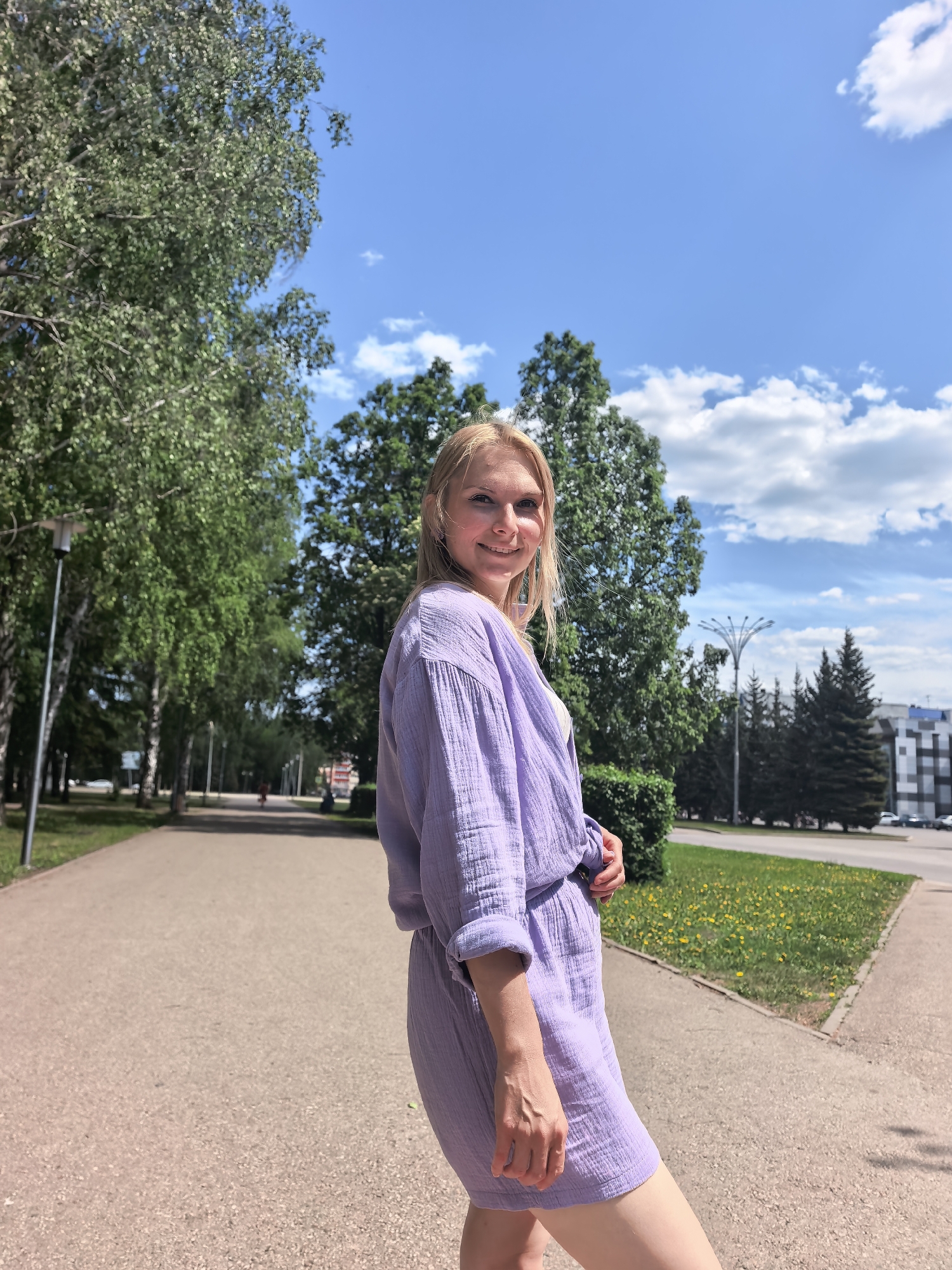 The photograph presents a woman standing on a paved walkway with trees lining the left side, while a grassy area dotted with yellow flowers is visible on the right. In the background, more trees are visible, and a building can be seen in the distance. The woman is wearing a light purple outfit. The sky is blue with scattered white clouds. The location appears to be a park or a similar public space. According to the metadata, the photograph was taken in the city of Sterlitamak, Russia.