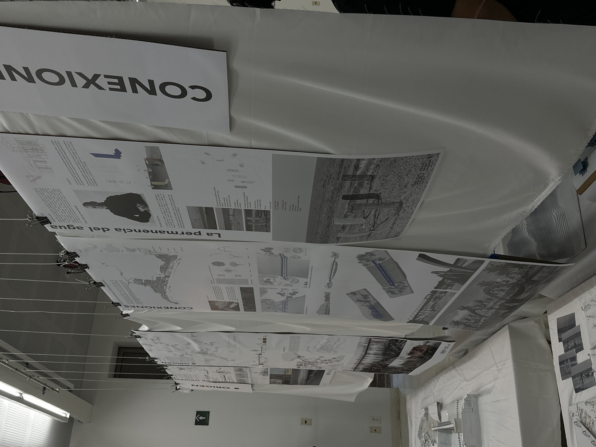This media file captures an indoor scene depicting an exhibition or presentation, likely in an academic or design context within Mexico City, Mexico. The primary subjects are numerous large-format posters or project boards, vertically suspended from a system of wires or strings using binder clips. These boards are layered, showcasing various architectural or design projects through technical drawings, conceptual diagrams, photographs, and descriptive text.
The activity taking place is an exhibition, where detailed design work is presented for viewing. On one of the upper posters, a portrait photograph of a man is visible, likely a student or a project participant. Below the hanging displays, a white-draped table holds additional project materials, possibly including architectural models or smaller sketches. The setting appears to be a well-lit interior space, with fluorescent lighting fixtures visible on the ceiling and an emergency exit sign on a wall in the background.
Notable details include the prominent use of Spanish text. A large banner at the top displays "CONEXIONES" (Connections). Other visible titles on the posters include "La permanencia del agua" (The permanence of water), "ORIGEN" (Origin), and "PUNTO DE ORIGEN" (Point of Origin). The word "IBERO" is also discernible on one poster, suggesting a connection to Universidad Iberoamericana, a known institution in Mexico City. The overall atmosphere points to a professional or educational event, possibly a critique or public display of student work.
