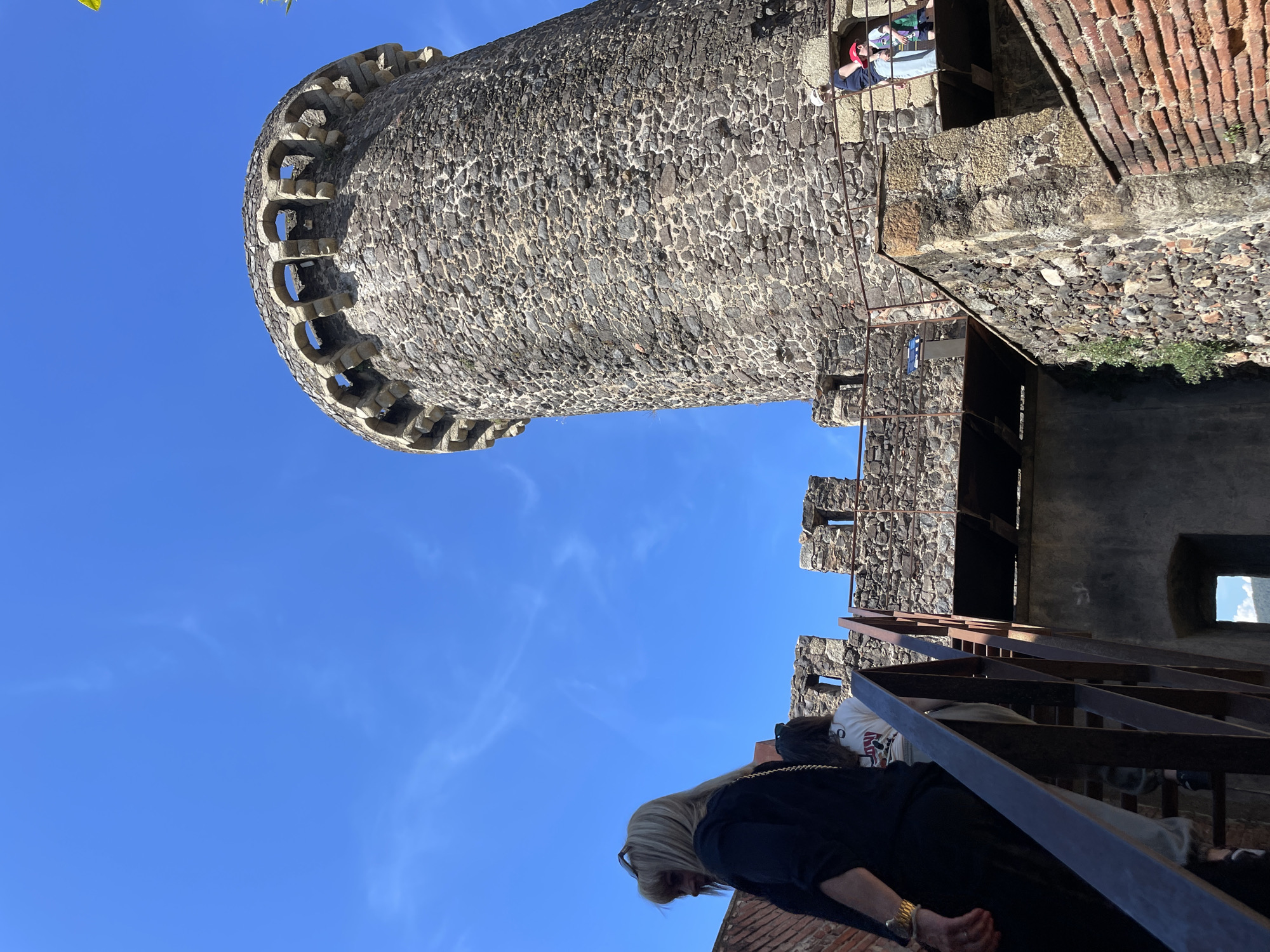 A low-angle shot reveals a medieval stone tower, likely part of a castle, set against a clear blue sky. The tower is made of rough, dark stones and features a crenellated top. Adjacent to the tower is a structure made of mixed stone and brick, with metal stairs leading up. Two people are visible in one of the tower's upper windows. At the bottom of the frame, a person with blonde hair and a dark blue garment is walking up the metal stairs.
This is a scene at the Castle of Hostalric, a medieval fortress in Hostalric, Spain.