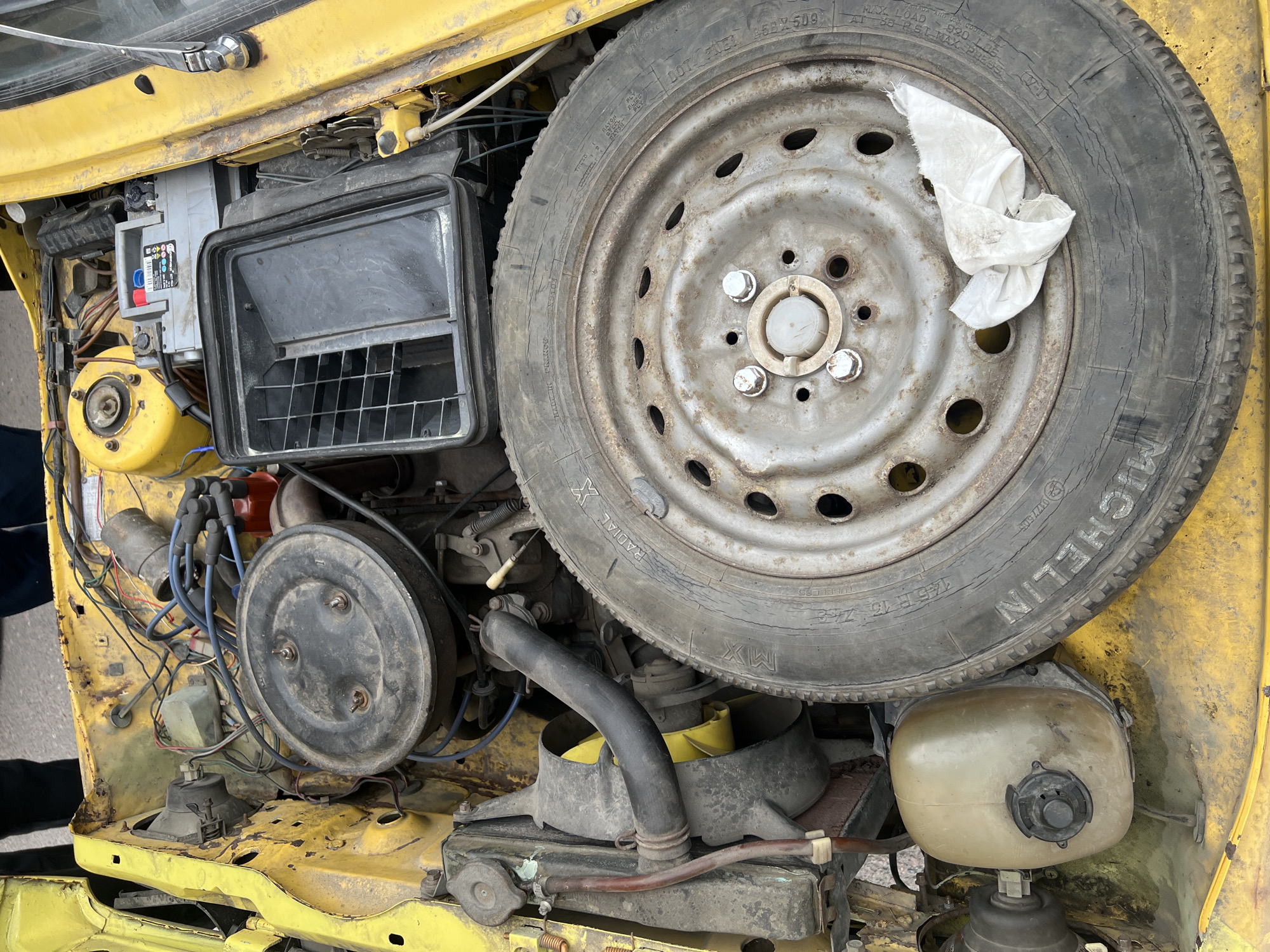 The image depicts the engine compartment of a yellow vehicle, possibly a Lada or other Soviet-era car. A spare tire is positioned on top of the engine, taking up a significant portion of the frame. The tire is a Michelin radial and exhibits signs of wear. The engine itself is visibly dirty and appears to be an older model, with various wires, hoses, and components exposed. A battery and air filter housing are visible on the left side of the engine.
The overall condition suggests the vehicle is either abandoned, undergoing repairs, or being stored. The lack of license plates or identifiable markings makes it difficult to determine the exact make and model of the car or its specific location. However, based on the vehicle type and general appearance, the scene could plausibly be situated in Saint Petersburg, Russia, or another location with a history of Soviet-era vehicles.