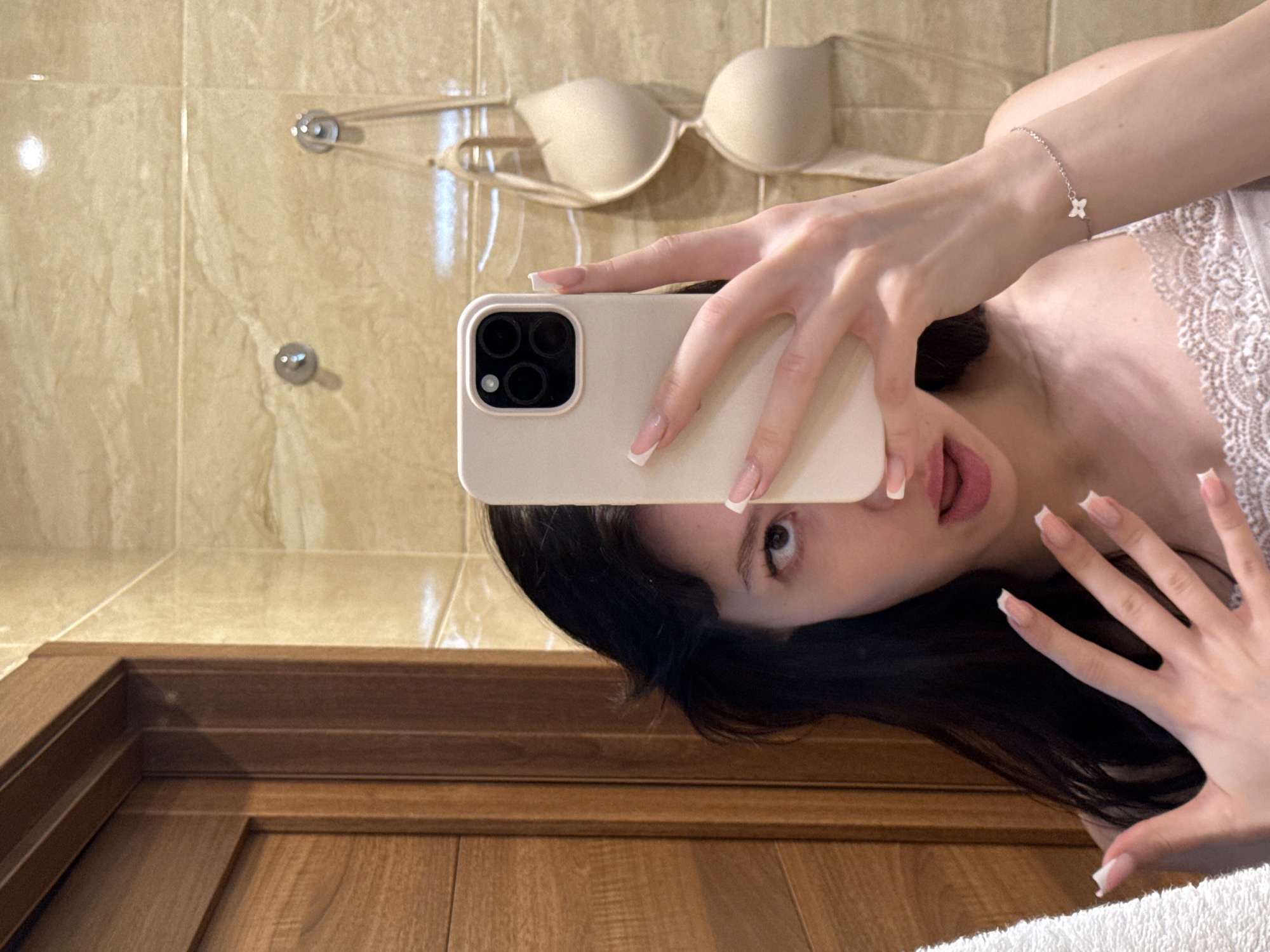 A young woman with long, dark hair and manicured nails is taking a selfie in a bathroom. She is wearing a white lace top and a silver bracelet with a small flower charm. In the background, a beige bra is hanging on a hook, and a wooden door is visible to the left. The bathroom walls are tiled with a marble-patterned material. The scene appears to be in an indoor residential setting, potentially a private home or hotel in Khimki, Russia.