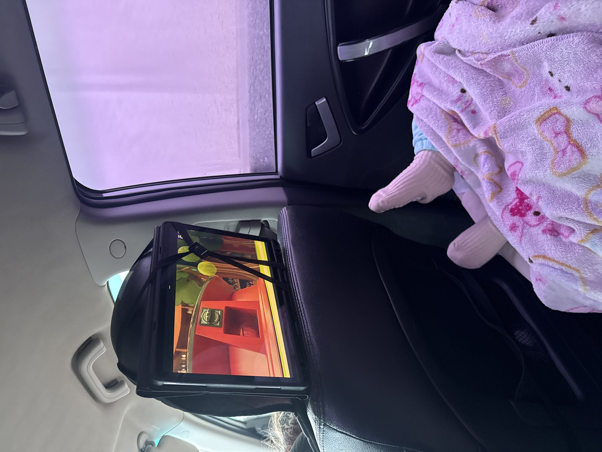 This media file captures an interior view of a car, looking towards the rear seat and out a sunroof or large window on the left. The scene appears to be set during the day, with bright, diffused light visible through the windows, casting a distinct purple hue, strongly suggesting the car is inside an automated car wash tunnel.
In the back seat, a child or infant is partially visible, covered by a light pink blanket with indistinct patterns. Only their feet, clad in matching light pink socks, are clearly seen. Mounted to the back of the front passenger seat headrest is a tablet, secured with black elastic straps. The tablet screen displays a vibrant, colorful cartoon or animated scene, featuring red structures, yellow circular objects, and other bright elements. The back of a person's head, likely an adult, is partially visible in the lower center-right, showing dark hair with some lighter strands and a patterned head covering or headband.
The car's interior features black leather seats and dark plastic trim, contrasted with lighter gray or beige headliner and grab handles. A car door with a silver handle accent is visible on the upper right. The overall impression is one of a comfortable journey, with the child entertained by the tablet during a car wash. The specific location, Kiselevka, Russia, is provided, though no visual cues within the image confirm this detail. There is no discernible text visible in the image.