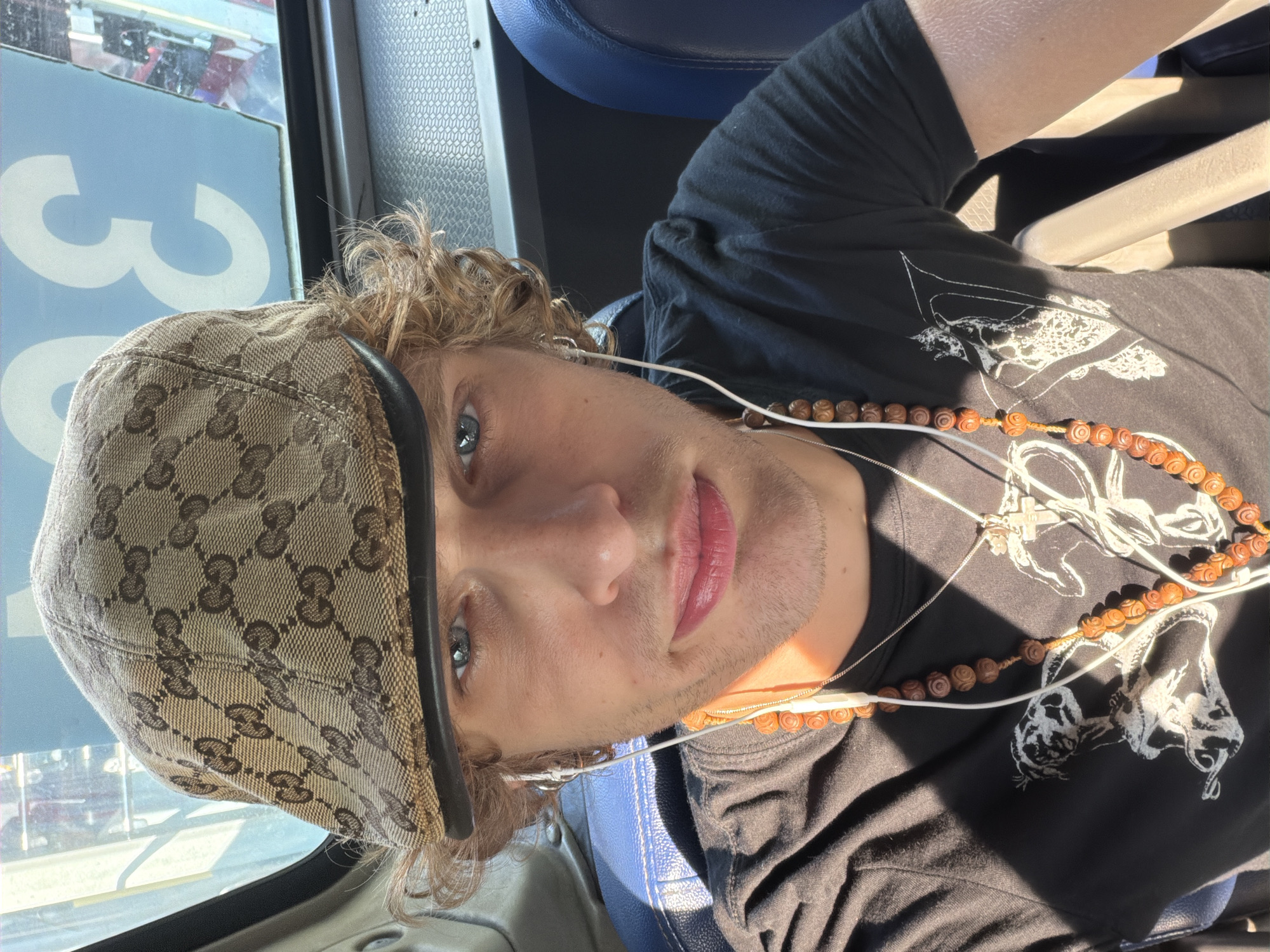 A young, fair-skinned male with curly blonde hair and blue eyes, wearing a beige Gucci monogrammed flat cap, white earphones, a black t-shirt with white graphics, and two necklaces, one with wooden beads and a silver cross pendant, is seen taking a selfie indoors, likely on public transportation. The background shows blue seats and a window with a partial blue sign displaying the number "30." The lighting suggests a sunny day. There is no indication of the location being in Seoul, South Korea.