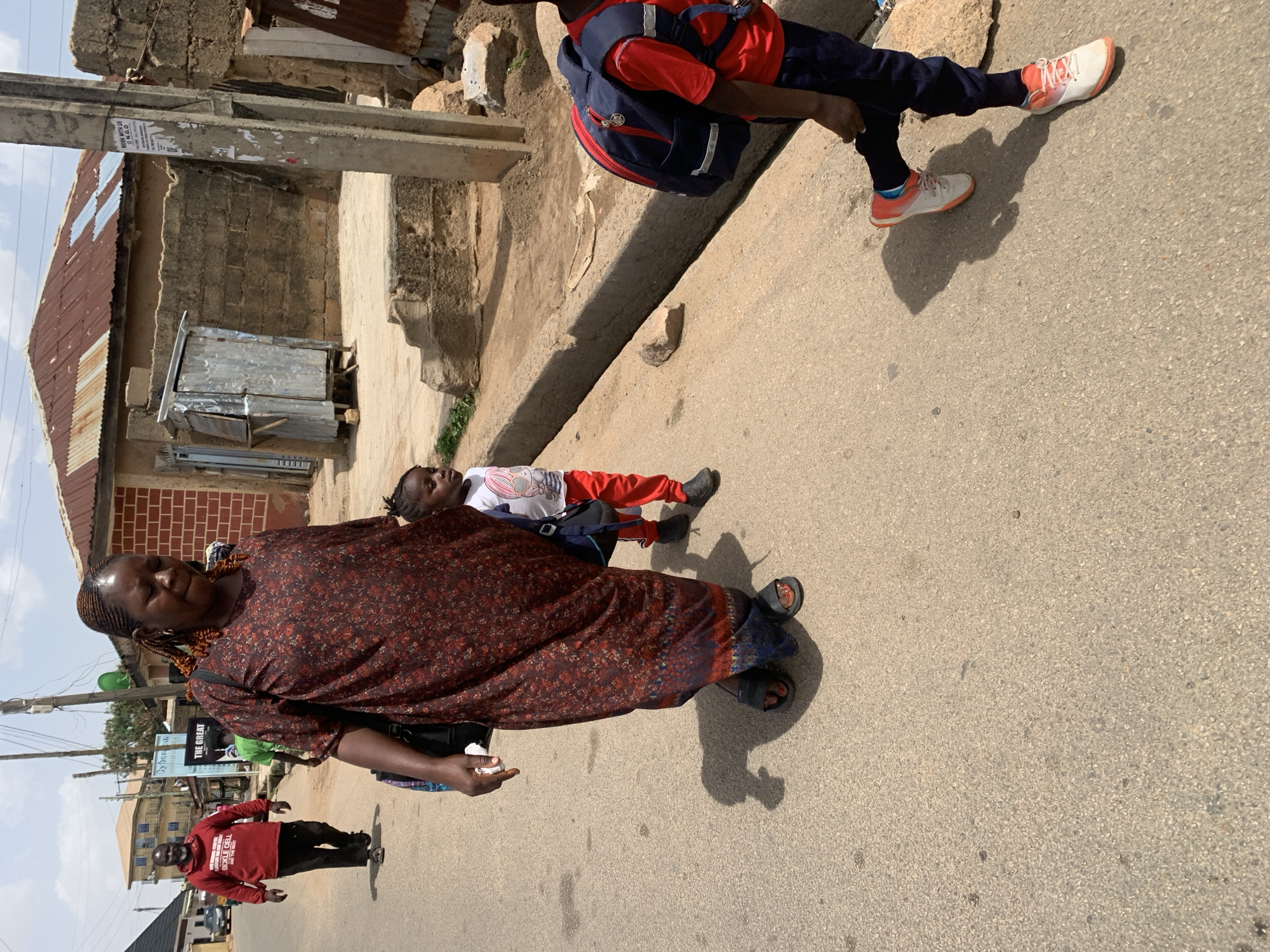 The image shows a street scene in Jos, Nigeria. A woman wearing a patterned brown dress and black sandals is walking toward the camera on a paved street. A younger person lies on the ground behind her, and another person wearing a red shirt, dark pants, and orange sneakers is sitting on a low concrete wall to the right. There is a metal shack and a brick building to the left of the street. A man in a red shirt and black pants is walking away from the camera further down the street. Overhead power lines and other buildings are visible in the background. The lighting suggests it is daytime.