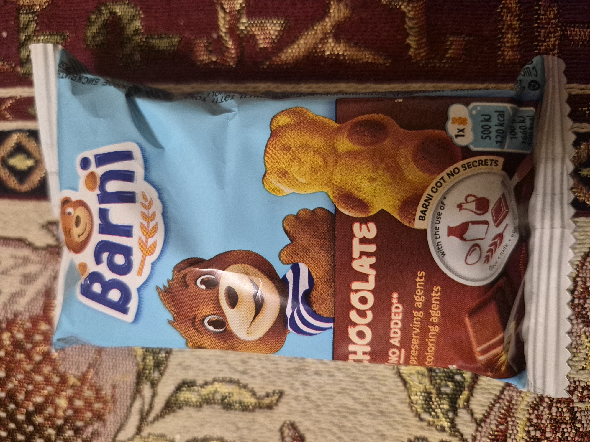 A Barni snack package, likely a soft cake, is prominently displayed horizontally on a patterned fabric surface, resembling a rug or tablecloth, in Yerevan, Armenia. The package design features a light blue background, with a rich brown section indicating the chocolate flavor. The brand's mascot, a friendly brown bear named Barni, is depicted wearing a blue and white striped shirt. A realistic image of the bear-shaped cake product is also visible on the packaging.
Visible text on the package includes "Barni" as the brand name and "CHOCOLATE" for the flavor. Key marketing points state "NO ADDED** preserving agents coloring agents" and "BARNI GOT NO SECRETS with the use of" accompanied by icons representing flour, milk, and eggs. Nutritional information is presented in a cloud-shaped graphic: "1x 500 kJ 120 kcal" per serving, and "100g 1660 kJ 400 kcal." A "KZ" logo, possibly for Kazakhstan standards, is also seen. Partial Cyrillic text, such as "TOKYONHOE 5NICKBN," is visible along the top edge. The image is a static product shot with no activity taking place, set against a blurred background of reddish-brown and beige patterns.