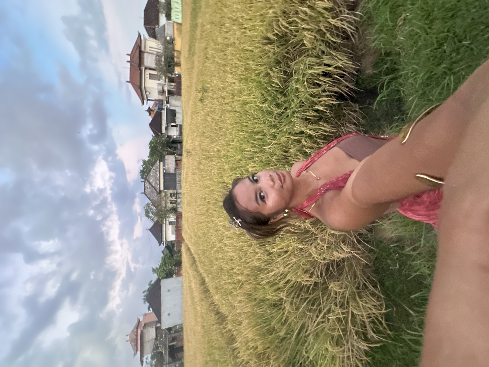 A person with tan skin and brown hair is positioned in a field of tall, golden grass. They are wearing a red dress and a gold necklace. In the background, there are houses with red roofs and white walls, set against a cloudy sky. The scene is captured from a close-up, low-angle perspective. The location is Sukawati, Indonesia.