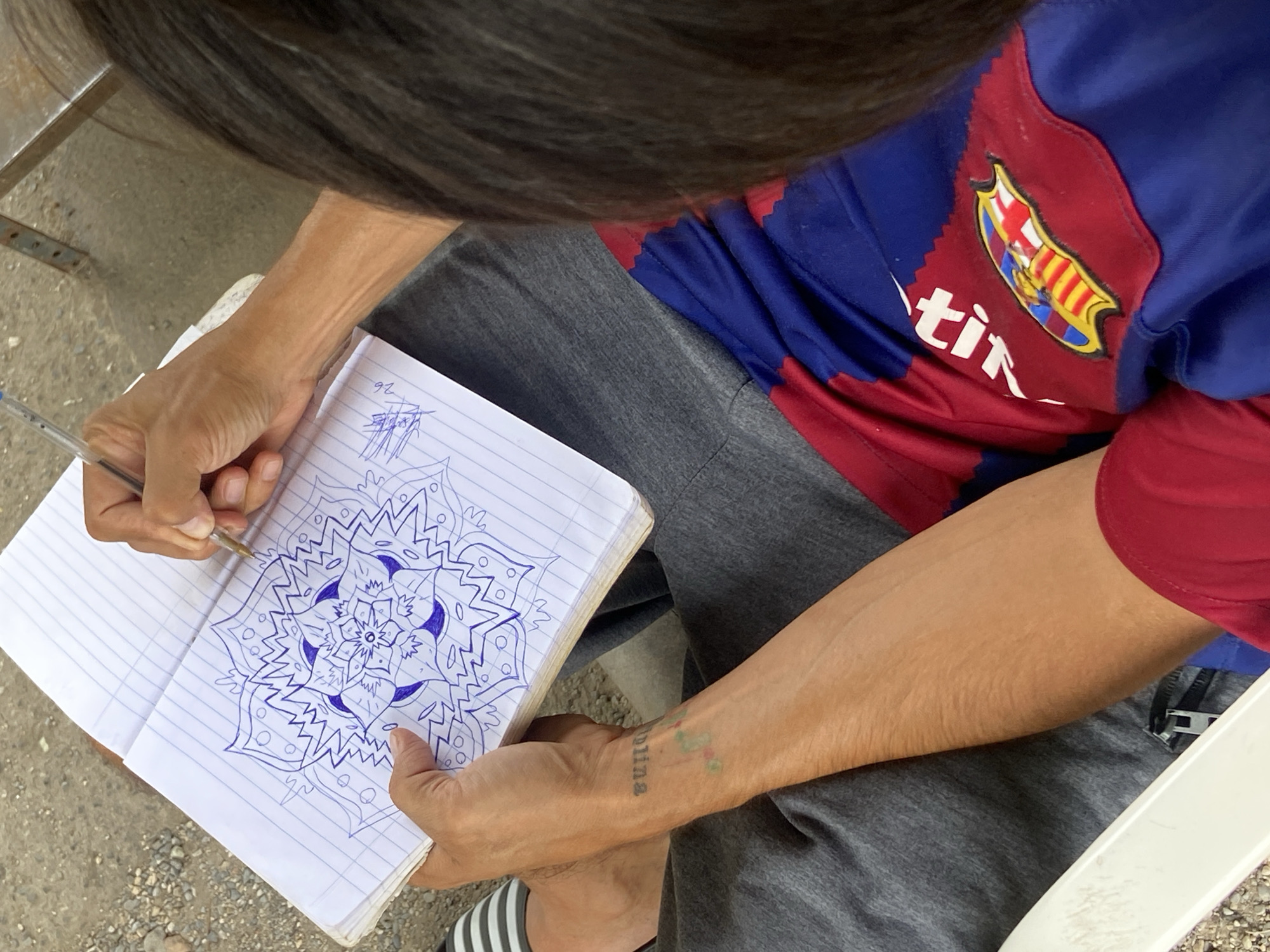 A person with dark hair is seated, wearing a Barcelona FC jersey (blue and maroon) and grey sweatpants. They are drawing a blue mandala-like design in a lined notebook. The person's left arm is visible, showing a tattoo that reads "tina." The location is Tinaquillo, Venezuela. The background is blurry, but appears to be an outdoor setting.