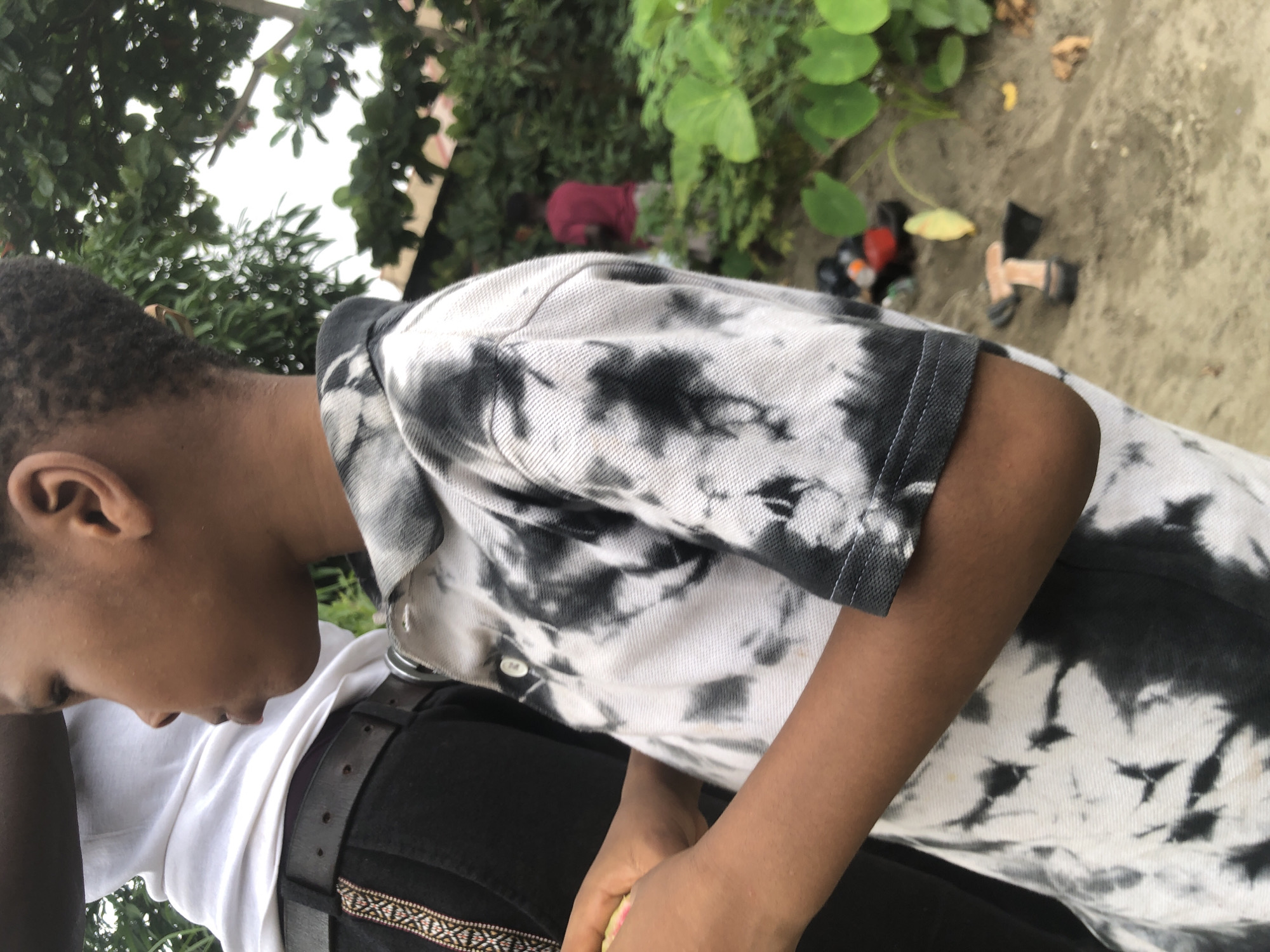 In Ado, Nigeria, this image captures a close-up of a young boy in the foreground, seen from the side profile. He is wearing a black and white tie-dye polo shirt. To his left, another person, partially visible, wears a white hooded garment and dark pants with a patterned belt. The background features lush green foliage and trees, suggesting an outdoor setting possibly near a beach or sandy area, indicated by the sand and some scattered objects like shoes and a yellow leaf on the ground. In the distance, another individual in a red shirt can be seen amidst the greenery. The lighting suggests it is daytime.