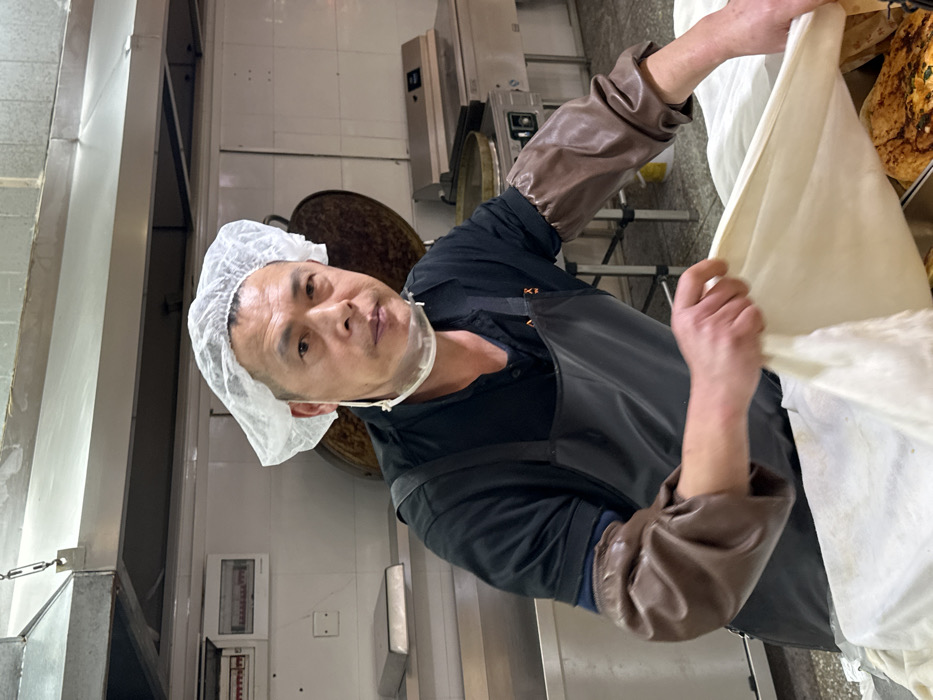 A male food handler is depicted within a commercial kitchen setting in Jiangjin, China. The individual, an adult male with short dark hair, directly faces the viewer with a neutral expression. He is attired in a white disposable hairnet, a clear plastic chin-level face shield, a black short-sleeved shirt, a dark apron, and brown protective sleeves covering his forearms. His hands are actively manipulating a large, light-colored sheet, consistent with dough or fabric, draped over a metal tray. A portion of browned, cooked food is visible beneath the sheet. The background features a stainless steel kitchen exhaust hood, white tiled walls, an electrical panel mounted on the wall to the left, a rectangular silver tray on a lower shelf, and various industrial kitchen appliances including a large circular dark pan on a stand and a stainless steel counter with an integrated cooking surface to the right.