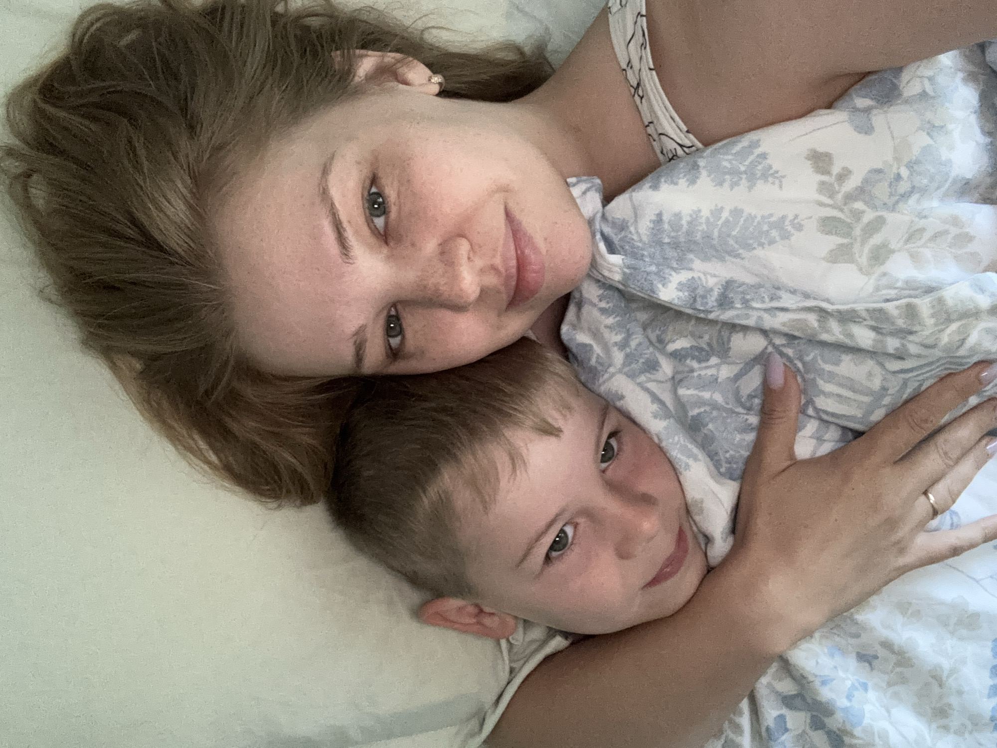A selfie shows a woman and a boy lying in bed, seemingly at home in Rusanovo, Russia. The woman, with her right arm around the boy, is positioned on the right side of the frame. She has light brown hair that is partially covering her face, light green eyes, and freckles. She is wearing a white top with a subtle pattern. The boy, on the left, has short brown hair, light green eyes, and fair skin. He is nestled into the woman's embrace, looking towards the camera with a slight smile. Both are covered by a light-colored blanket with a blue floral pattern. The overall impression is one of intimacy and comfort, suggesting a moment of shared rest or affection.