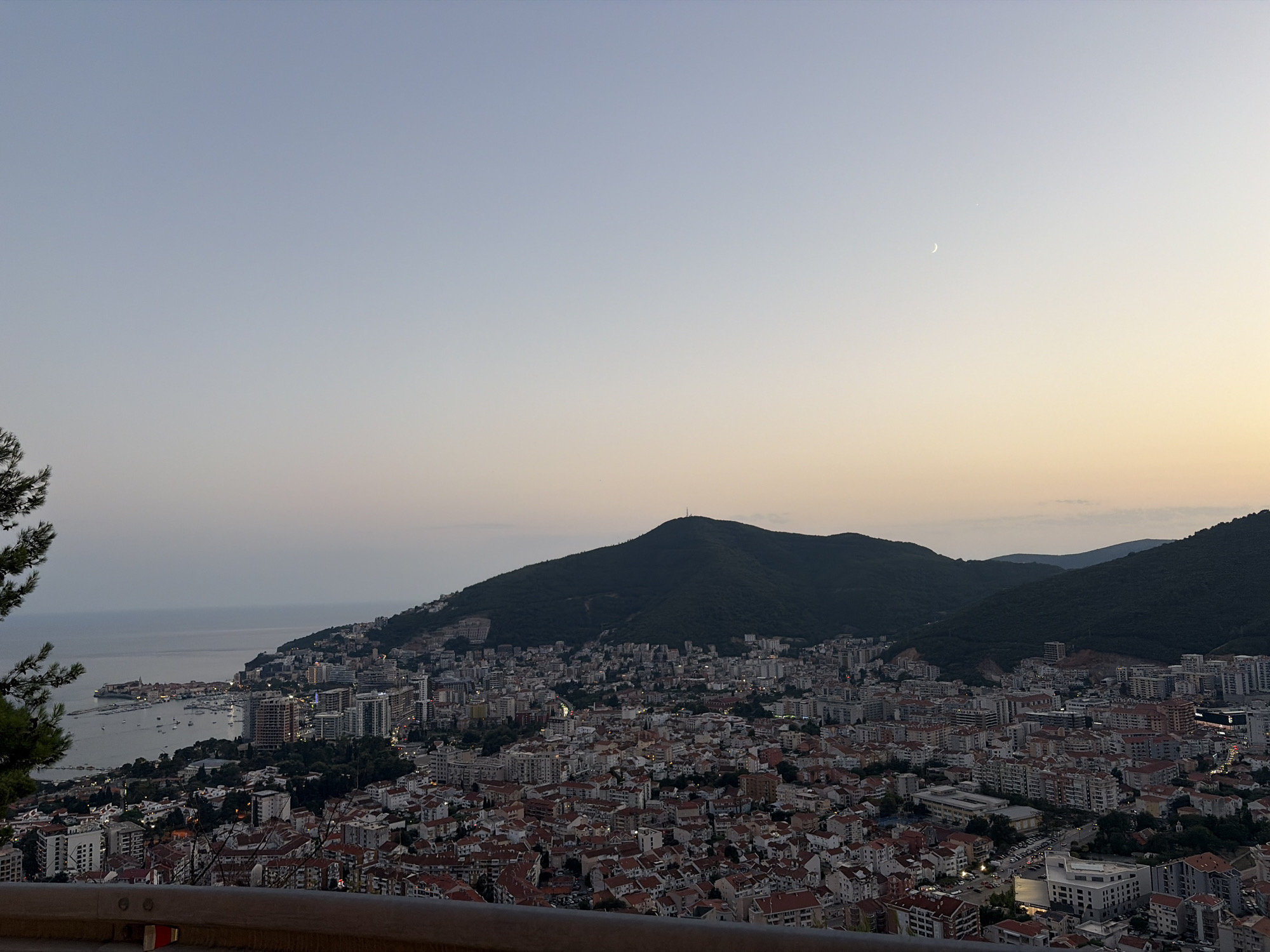 An elevated panoramic view captures the coastal city of Budva, Montenegro, nestled against a backdrop of lush, dark green mountains. The sky transitions from a pale blue at the top to a soft, warm gradient of yellow and orange near the horizon, indicating either sunrise or sunset. A faint crescent moon is visible in the upper right portion of the sky, accompanied by a small, bright star.
The city sprawls across the hills, a dense collection of buildings with red-tiled roofs interspersed with taller, modern structures. Lights are beginning to illuminate the city, casting a glow on the urban landscape. Along the coast, a bay is filled with numerous boats and yachts, their lights reflecting on the calm water. The shoreline is a mix of urban development and natural terrain.
In the foreground, a dark metal railing runs horizontally across the bottom of the frame, suggesting the viewpoint is from a road or a raised platform. To the left, the dark silhouette of pine trees partially obscures the view of the city, framing the scene. The overall impression is one of a tranquil evening over a bustling coastal city.