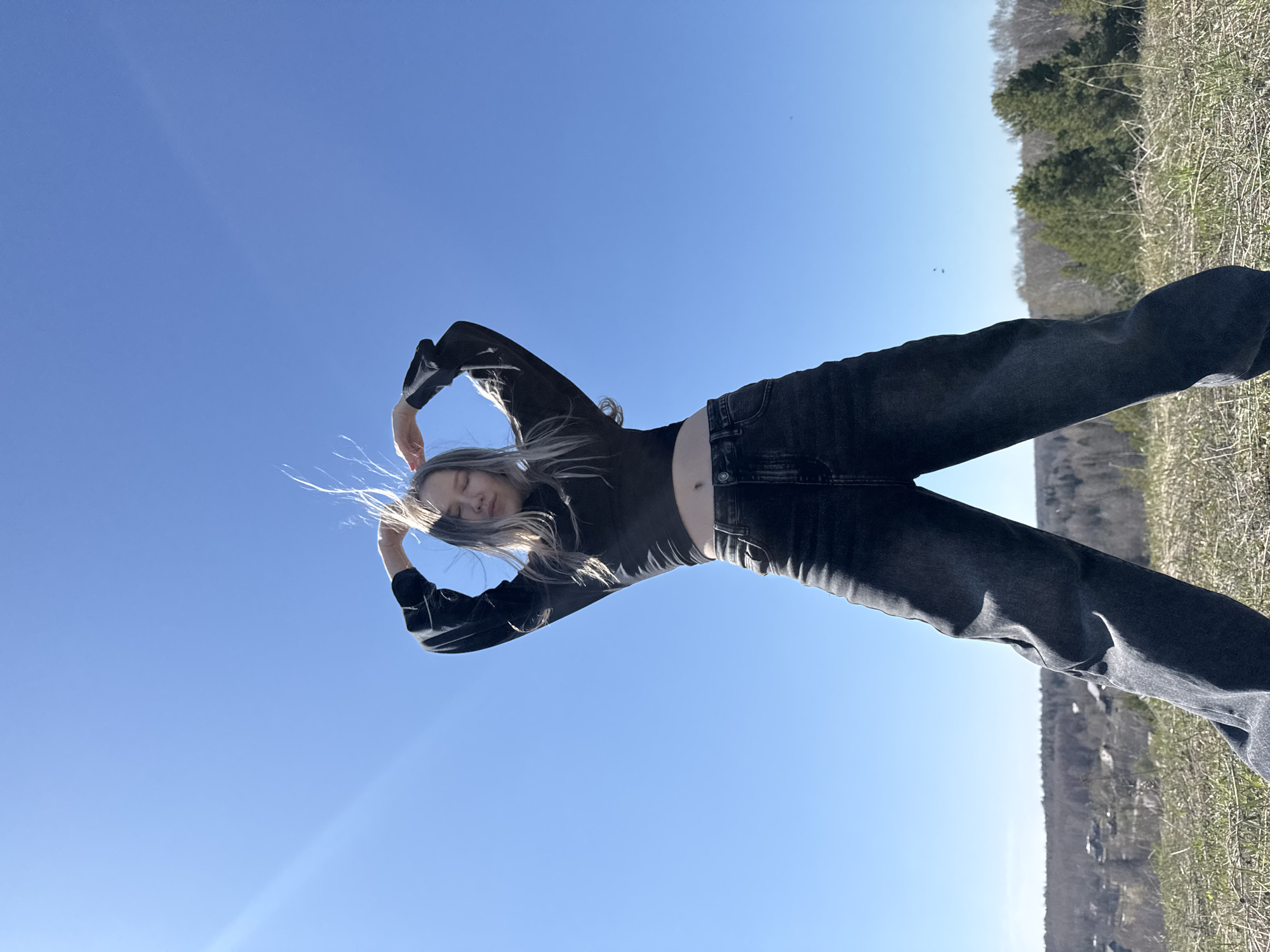 A slender person is depicted leaning backward in a landscape. The sky is a clear blue. The person has long, gray-white hair and wears a black long-sleeved top and dark-colored jeans. Their arms are positioned above their head, forming a slight curve. The background shows dry grass and trees in the distance.
The image appears to have been taken at ground level, looking up at the subject. The location is Zhigalki, Russia.