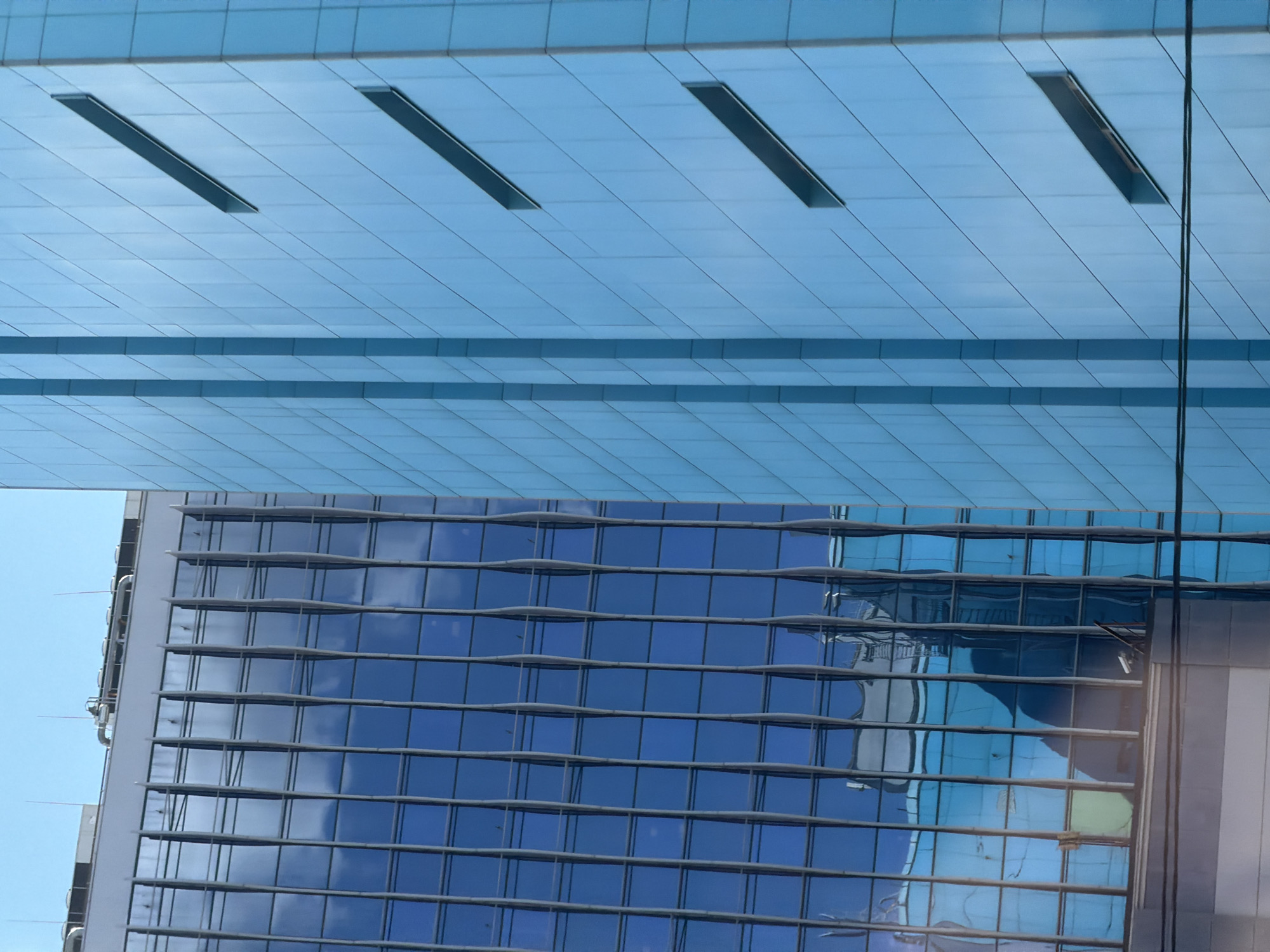 This media file captures an abstract, angled view of modern architecture, likely in a city setting. The primary subjects are two large building facades, both featuring extensive use of blue glass.
The upper portion of the image shows a light blue, tiled ceiling or facade with subtle horizontal lines and three dark, rectangular recessed openings. This area appears to be a large, flat plane.
Below this, the image transitions to a darker blue, glass-clad building. This building's facade is characterized by a grid of horizontal and vertical lines, suggesting a windowed structure. The glass reflects the sky, appearing as patches of bright blue interspersed with darker, cloud-like shapes. The reflections also hint at the presence of other buildings or architectural elements, creating a layered and somewhat distorted visual effect. The reflections are fluid and abstract, adding a sense of depth and movement to the otherwise static structures.
There are no visible people or explicit activities taking place. The scene is dominated by the geometric shapes and reflective surfaces of the architecture. The lighting suggests daylight, with clear blue sky visible in the reflections and the background, indicating fair weather. The overall impression is one of urban scale and contemporary design, with a focus on the interplay of light, reflection, and geometric form. No text is visible in the image. Given the location context of Moscow, Russia, these buildings are likely part of the city's modern skyline.